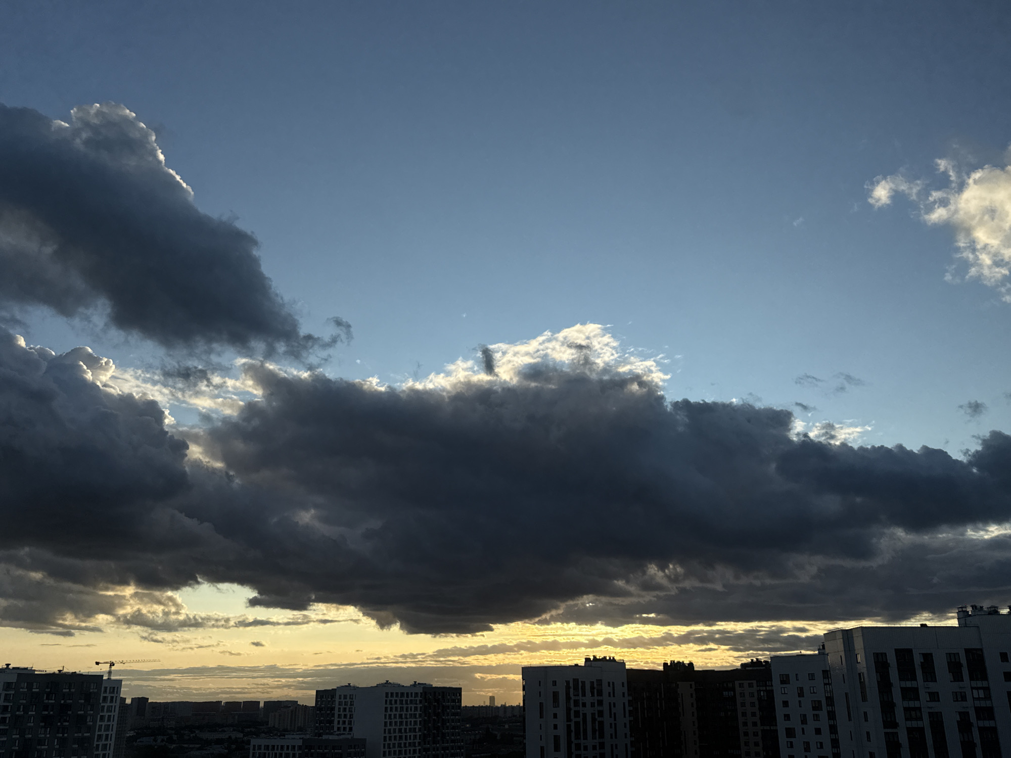 The image depicts a sky over the city of Lyubertsy, Russia, during what appears to be late afternoon or early evening. Dark, dramatic clouds dominate the upper portion of the frame, with some illuminated edges suggesting the setting sun behind them. In the distance, a hazy horizon with more clouds can be seen. The lower part of the image shows a collection of residential buildings, some appearing to be modern high-rises. A construction crane is visible in the far distance, indicating ongoing development. The sky is a mix of deep blues and greys, with streaks of yellow and orange near the horizon, characteristic of a sunset or sunrise. There are no people visible in the image.