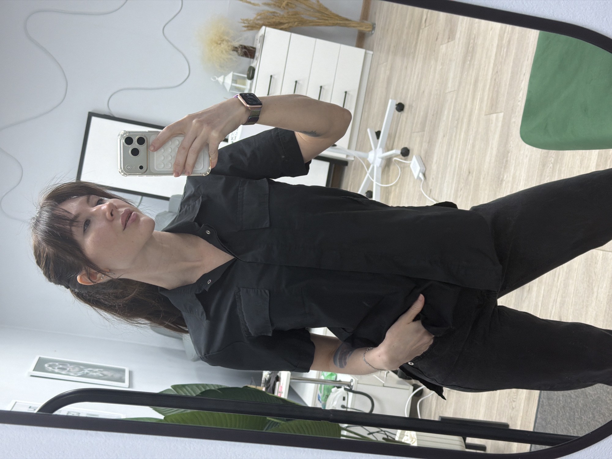 A woman stands in front of a mirror, taking a selfie with a smartphone. She is wearing a black short-sleeved shirt and black pants. The woman has brown hair with bangs. She is wearing a watch on her left wrist. Behind her is a white dresser with decorative items on top. A green chair is visible in the background. The location is identifiable as Perm, Russia due to the interior design.