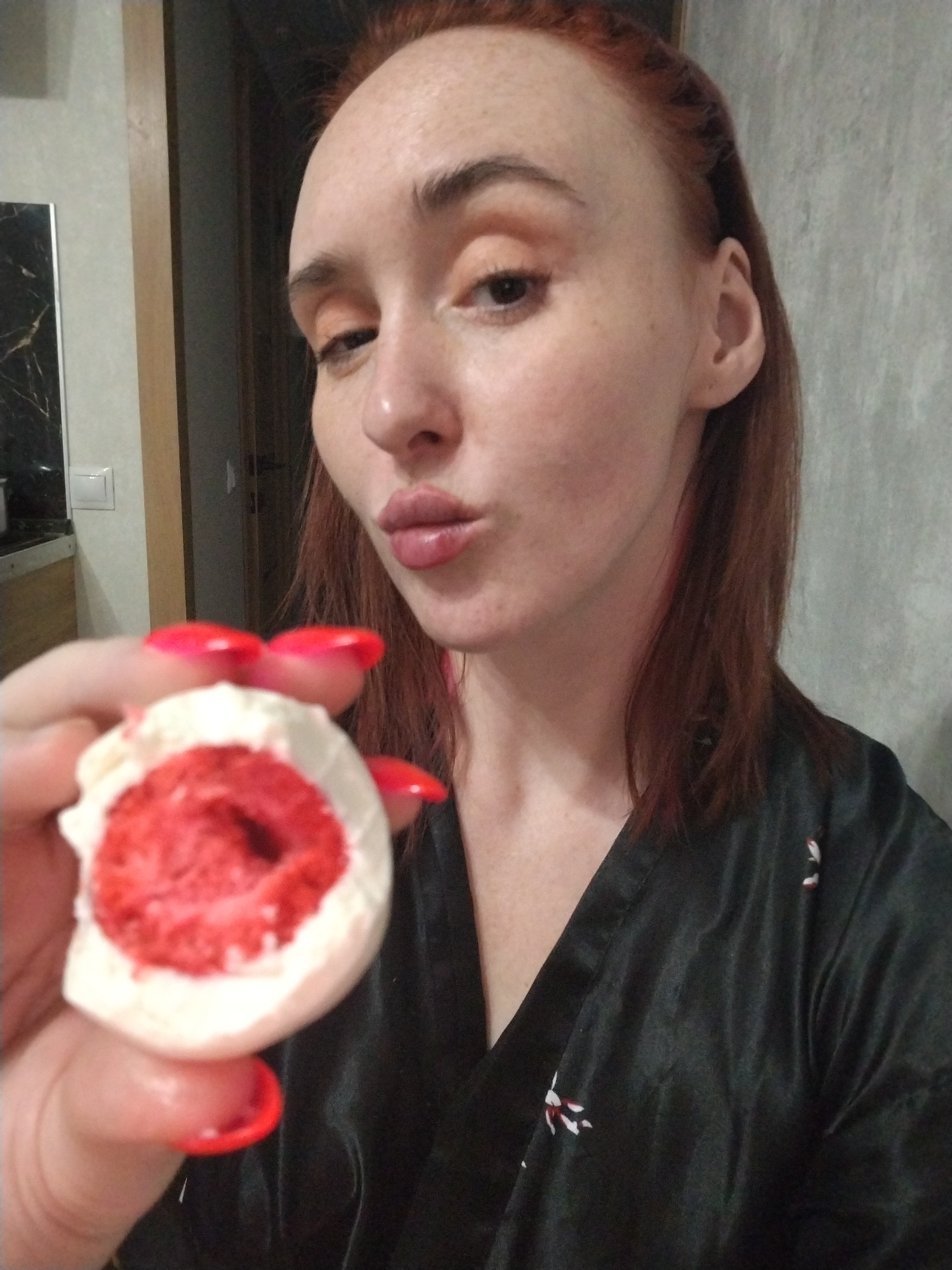 A woman with red hair and freckles is holding a bitten dessert with red filling and white frosting, positioned near her face. She has glossy lips, and her nails are painted bright red. She is wearing a black robe with a floral pattern. The background includes a gray wall and a glimpse of a kitchen area. The location is not discernible from the image.