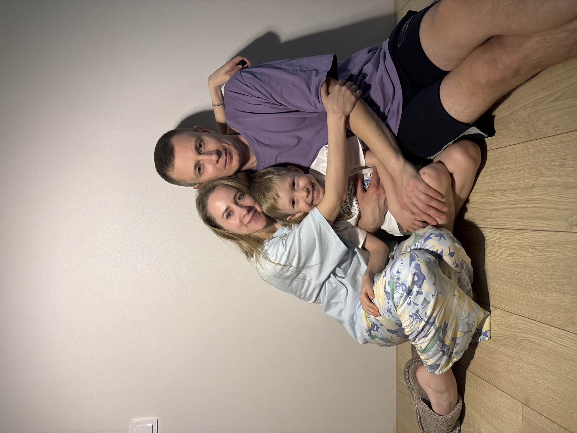 The image captures a heartwarming moment of a family of three – an adult man, an adult woman, and a young boy – lying on a light-colored wooden floor, leaning against a plain white wall. The scene is set indoors, likely in a home, with a minimalist aesthetic.
The man, sporting short dark hair, is positioned at the back, wearing a purple t-shirt and black shorts. His arms are wrapped around the woman and child, and he smiles gently towards the camera. In front of him is the woman, with long blonde hair, dressed in a light blue t-shirt and colorful floral-patterned pajama pants in hues of blue, yellow, and green. She also smiles, her face close to the child's, with a silver bracelet visible on her left wrist as her arm extends around the man. The young boy, with light brown hair, is nestled comfortably between the two adults, wearing a white t-shirt and displaying a wide, joyful smile directly at the camera. All three subjects exude happiness and contentment, with their expressions conveying love and comfort within their embrace.
Notable details include the smooth, light-colored wall and the light wooden floor. A white electrical outlet or light switch is partially visible on the lower left side of the wall. The woman wears a single fuzzy grey slipper on her right foot. The lighting is soft, casting gentle shadows on the wall behind the family, suggesting an intimate and relaxed atmosphere. No visible text is present in the image. The location context for this tender family moment is Chaikovsky, Russia.