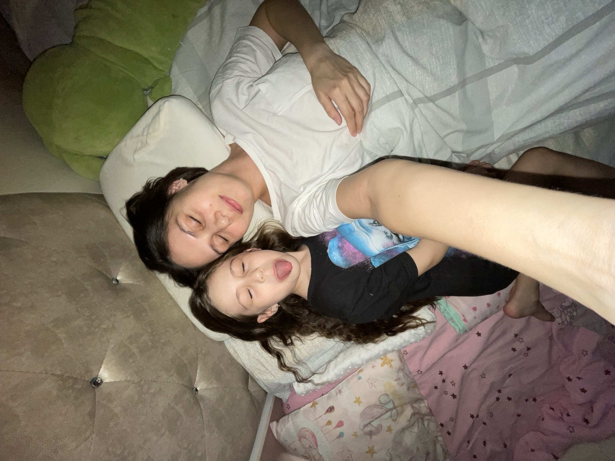 A top-down, slightly angled indoor shot depicts a woman and a young girl lying on a bed. The woman, with dark hair, wears a white T-shirt. Her eyes are closed. The girl, with long, curly brown hair, wears a black T-shirt with a graphic print and has her tongue slightly out. Both are resting on white pillows, which are on a grey velvet headboard with button accents. A large green stuffed animal is in the upper left corner. A grey comforter covers the woman. The girl is on a pink sheet with star patterns, resting atop several pillows with cartoon designs. A partial view of a person's arm and leg is in the upper right, suggesting someone is taking the photo. The lighting is dim. There is no information about the location of the scene in the city of Izhevsk, Russia.