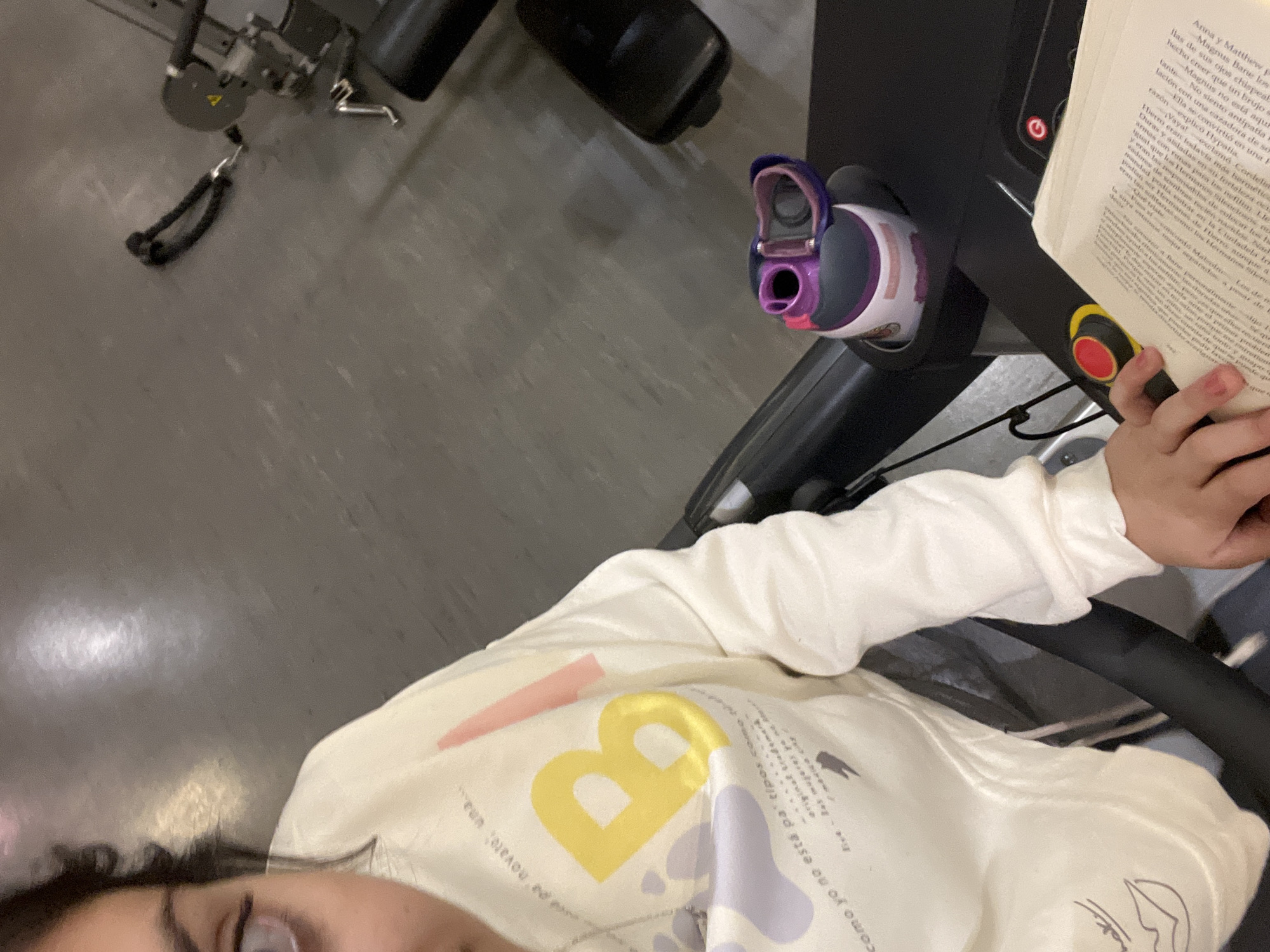 A person is on a treadmill, reading a book in Spanish. A water bottle sits in a holder on the treadmill. The person is wearing a white sweatshirt with a large yellow "B" on the front. Gym equipment is visible in the background, suggesting the location is a fitness center.