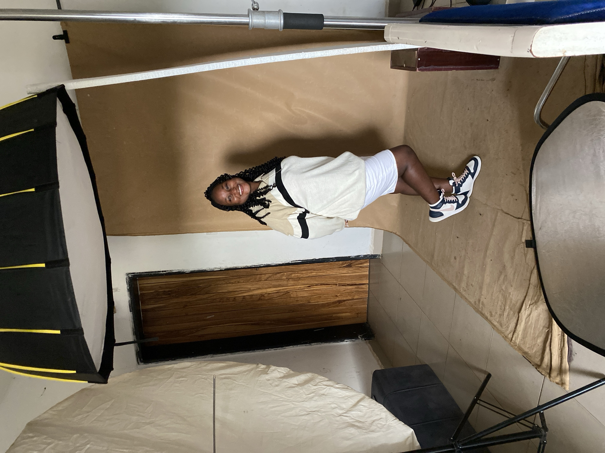 A woman wearing sneakers, a white skirt, and a tan and white striped sweater is laying on her side in front of a brown backdrop. The scene appears to be inside a photography studio, with a softbox on the left side of the image and a reflector on the right. A window with wooden slats is visible behind the woman. Based on image metadata, the location is Jos, Nigeria.