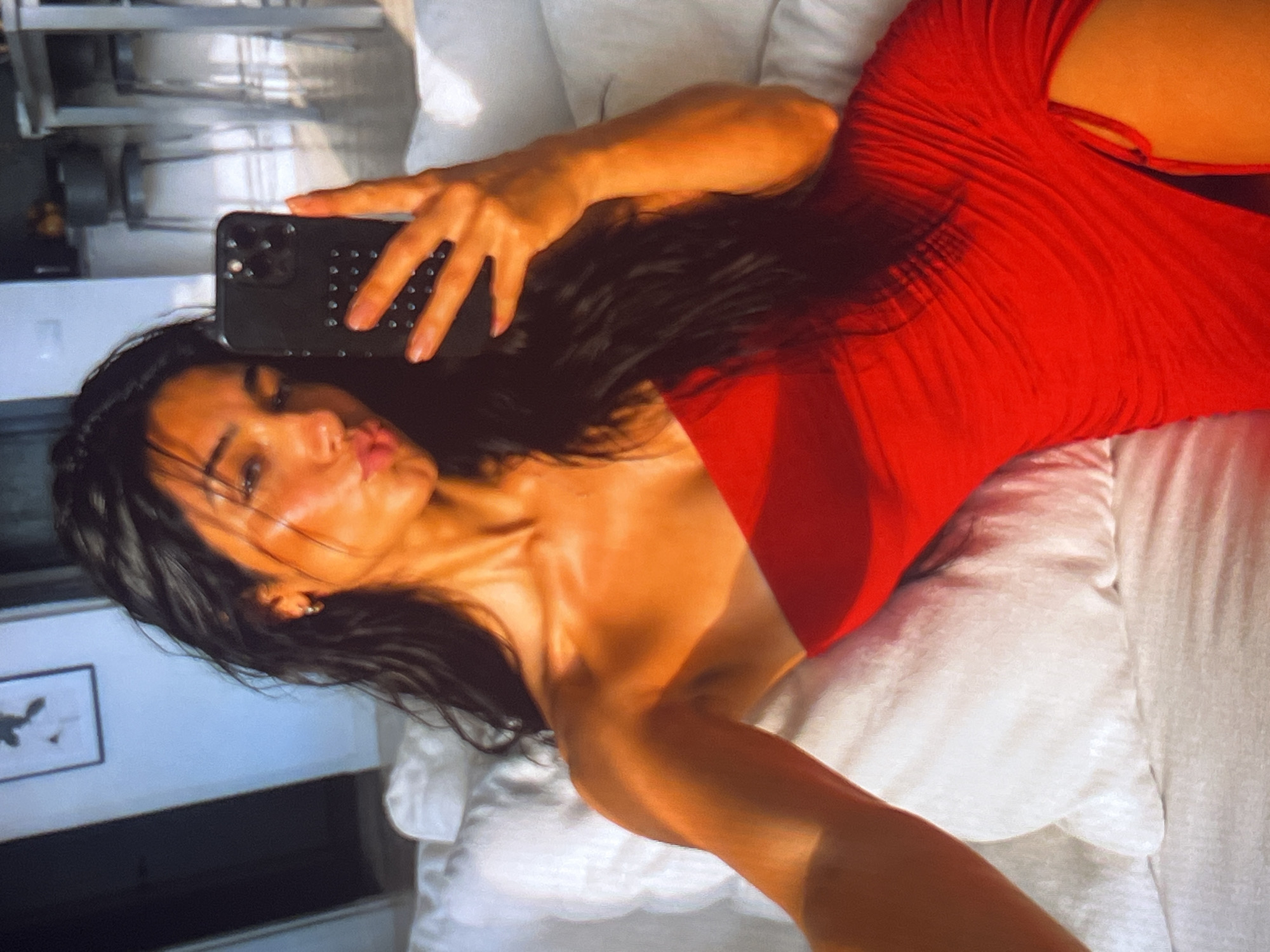 A young woman with long, dark, wavy hair, wearing a strapless red dress with a thigh-high slit, is taking a selfie in what appears to be a living room. She is seated on a white couch with plush cushions, holding up a black smartphone to capture the reflection. The lighting in the room is warm, casting a golden hue on her skin. In the background, a framed picture hangs on a light-colored wall, and further back, kitchen stools and a counter are visible. The woman is making a puckered-lip expression for the camera.