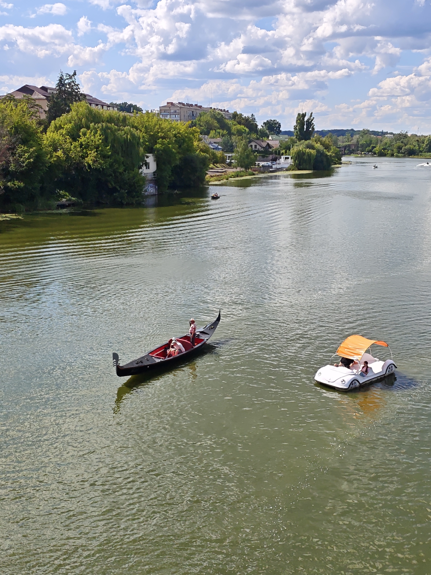 The image depicts a wide, calm river in Vinnytsia, Ukraine, with a lush, tree-lined bank visible in the background. The water has a greenish hue and is rippled by the movement of two recreational watercraft.
In the foreground, a traditional black gondola is being propelled by a person standing at the stern, dressed in a red and white striped shirt and white shorts. Two passengers are seated within the gondola, appearing relaxed. To the right of the gondola, a white pedal boat, shaped like a vintage car with an orange canopy, carries two occupants. One person is at the helm, steering, while another passenger sits beside them.
The riverbanks are densely populated with mature trees, their vibrant green foliage creating a natural border along the water. Behind the trees, hints of urban structures, including buildings with tiled roofs and apartment blocks, can be seen, suggesting the presence of the city. The sky is a bright blue, adorned with fluffy white clouds, indicating fair weather. The overall scene conveys a sense of leisure and recreational activity on the water.