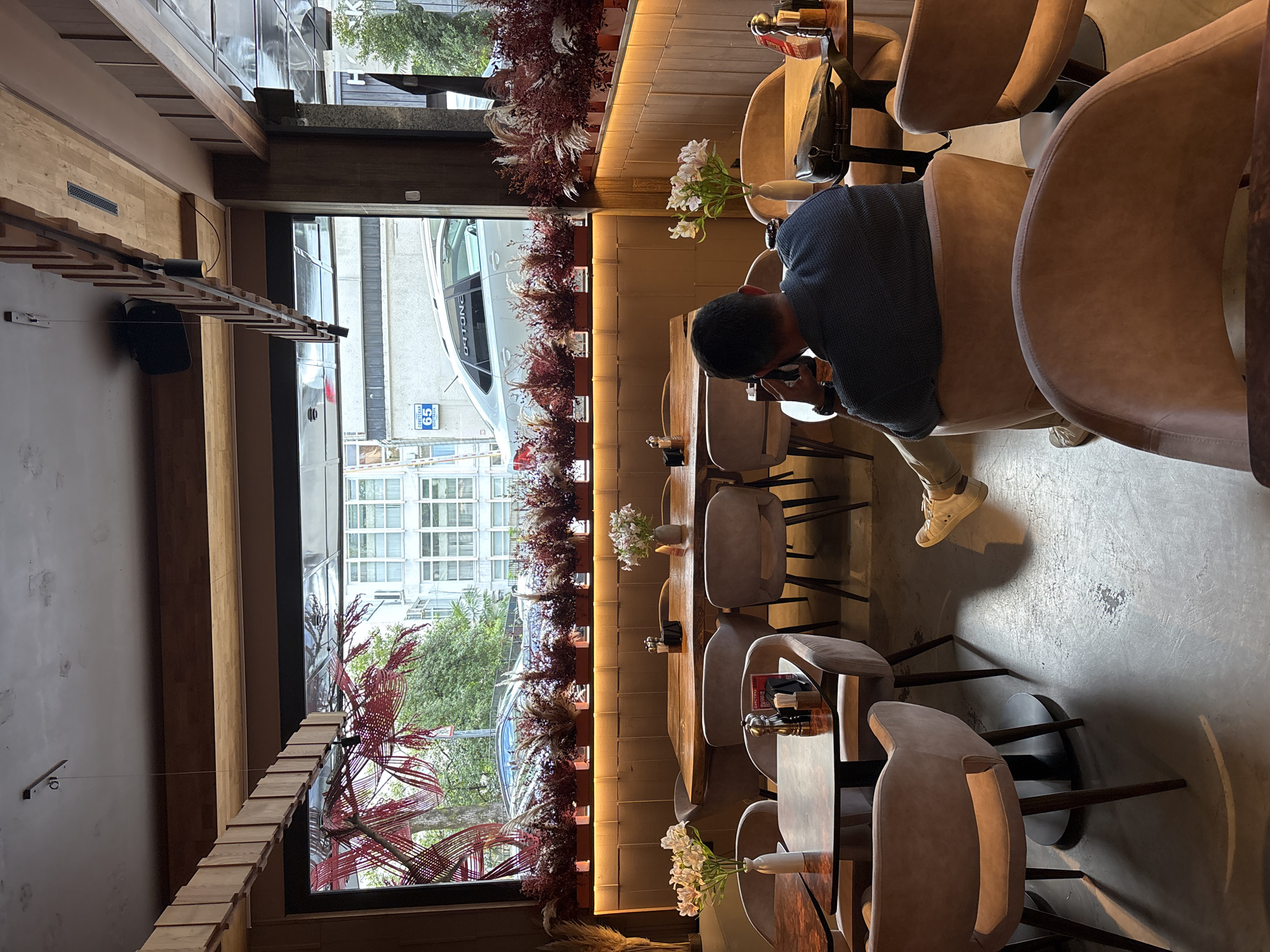 The image depicts the interior of a contemporary restaurant or cafe, likely located in Sochi, Russia, given the Cyrillic street sign visible through the window. The setting is characterized by warm, earthy tones, with wooden accents on the ceiling and walls, and plush, taupe-colored seating. A large window offers a view of a street scene, including buildings, trees, and passing vehicles.
In the foreground, a man is seated at a table, facing away from the camera, and appears to be engaged in a phone conversation. He is wearing a dark blue sweater and light-colored pants. In front of him, on the table, are a phone and a bag. The table also has salt and pepper shakers, and a napkin dispenser.
The restaurant's decor includes decorative plants and floral arrangements displayed on shelves and tables. The lighting appears to be a mix of overhead fixtures and ambient lighting from behind the wall panels, creating a cozy atmosphere. The floor is a polished concrete surface.
Visible through the window is a white sedan with a Cyrillic license plate and text that reads "СОЕТСКАЯ 65" (Sovetskaya 65), confirming the location. The window is further adorned with a red, intricate decorative element resembling a stylized tree branch or art installation. The overall impression is one of a modern, well-appointed establishment.