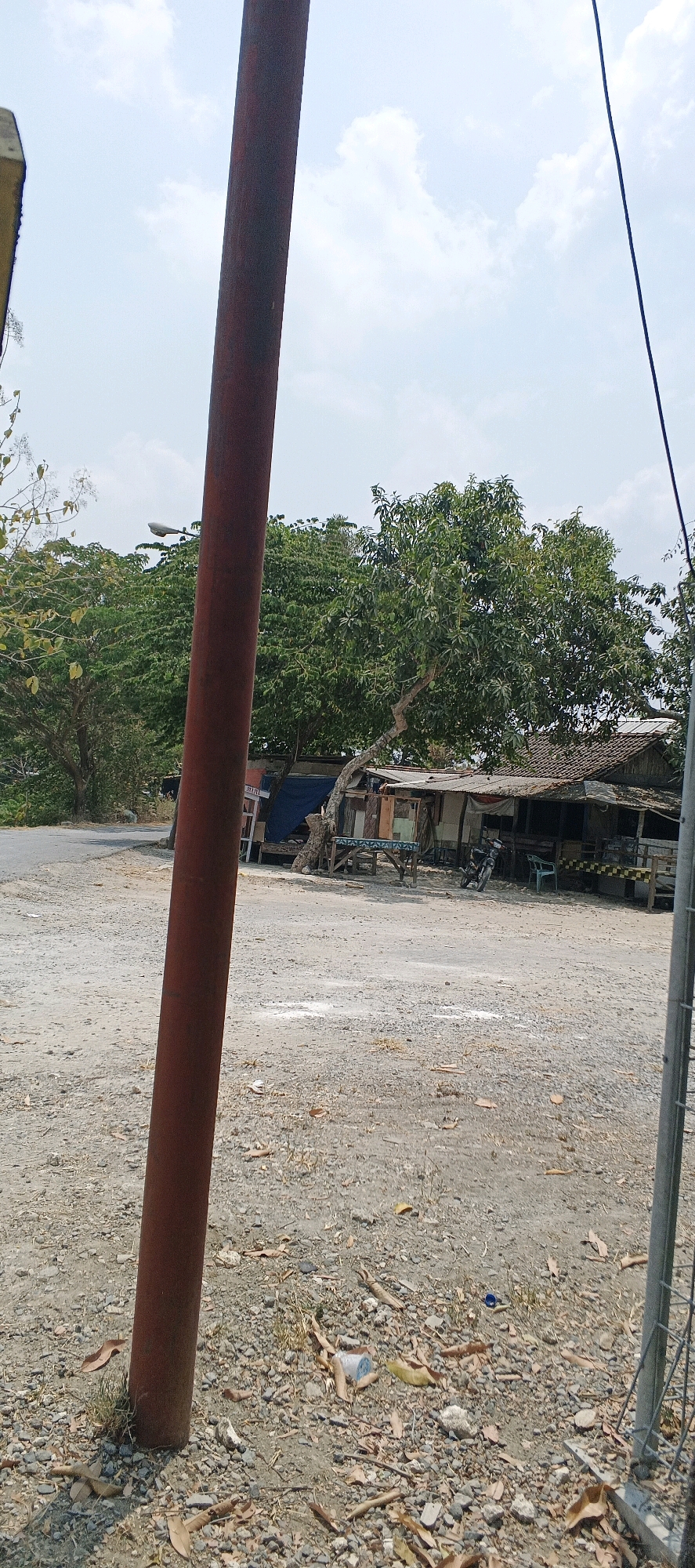The image depicts an outdoor scene in Parengan, Indonesia, featuring a gravel lot in the foreground and a collection of buildings and vegetation in the background. A prominent, rusty red pole is positioned vertically on the left side of the frame, partially obscuring a cloudy sky. The ground is covered with small stones, scattered dry leaves, and some debris, including a blue plastic bottle cap and a crumpled white and blue container.
In the midground, a group of structures, appearing to be simple shops or dwellings with corrugated metal roofs, are situated. One structure is notably leaning. In front of these buildings, there is a motorbike parked. A light-colored plastic chair is visible to the right of the motorbike. Behind the structures, a dense canopy of green trees dominates the background. A paved road is visible to the far left, with a streetlight mounted on a pole above it. The overall impression is of a rural or semi-rural area with modest infrastructure and abundant natural growth. There are no people visible in the image.