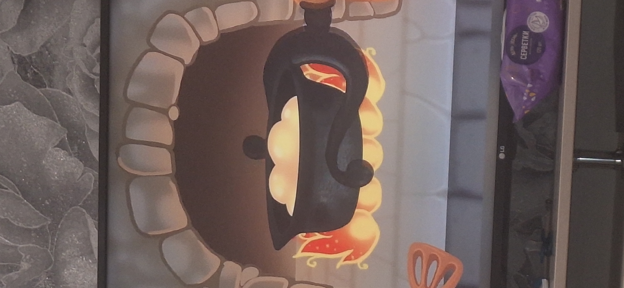 The image shows a cartoonish depiction of a black cauldron filled with light-colored, fluffy contents, sitting over stylized flames within an arched stone opening, resembling a hearth or oven. The cauldron has a lid with a small knob. Flames lick up from beneath the cauldron, some with red accents. To the left of the cauldron, a bright orange, curved object with three indentations is visible, possibly a decorative element or food item. The background of the hearth area is a dark brown. The scene is rendered in a flat, illustrative style. Below the hearth, the top of a dark electronic device, possibly a television, is visible. On the device, a white LG logo is present on the right side. To the right of the logo, a purple package with white text, including the word "СЕРВЕТКИ" (wipes), is partially visible. The image is likely a still from a video game or animation. The setting does not provide any specific clues to indicate it is located in Bila Tserkva, Ukraine.
