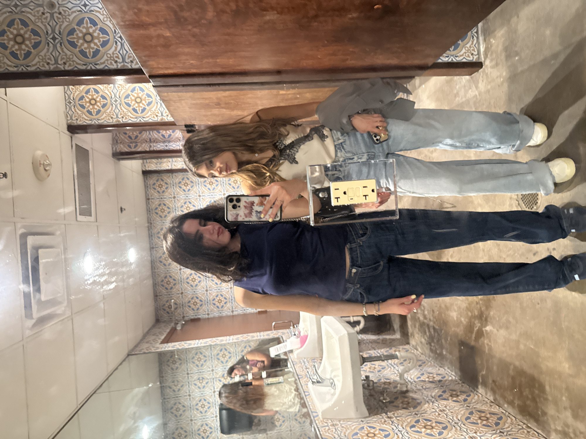 Two women are taking a selfie in a public restroom. The restroom has white tile walls and colorful tiled floors. There are white sinks visible in the mirror's reflection. One woman is wearing a dark blue shirt and dark blue jeans. The other woman is wearing a white top with a lace detail, light blue jeans, and white shoes. They are holding their phones, and one phone has a clear case with an electrical outlet attached to it. The location is College Station, United States of America.