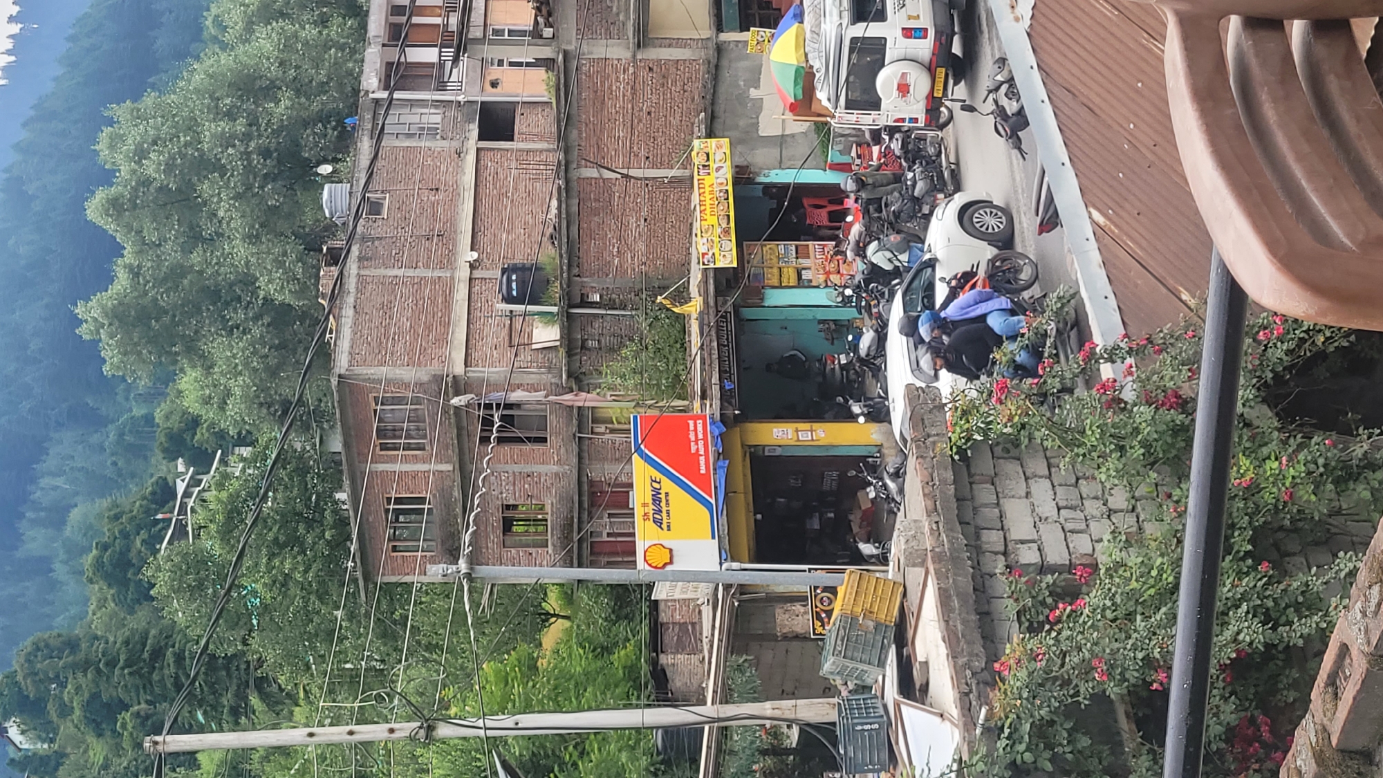The image depicts a street scene in Vashisht, India, characterized by a dense urban environment with multi-story brick buildings and a lush, forested hillside in the background. The foreground features a stone wall covered in blooming pink roses, with a black metal railing partially obscuring the view.
The street is bustling with activity. Several motorcycles are parked and in motion, with riders often wearing helmets. A white vehicle, possibly a van or SUV, is visible with a license plate. Adjacent to the street are various shops and establishments. One prominent sign reads "Shell Advance Bike Care Center," indicating a service center. Another sign displays "PAHADI DHABA," suggesting a local eatery. Crates and merchandise are visible outside some of the shops.
The architecture is typical of the region, with exposed brickwork and concrete structures. Power lines crisscross overhead, and water tanks are visible on the rooftops. The overall impression is one of a lively, commercial street in a mountainous area.