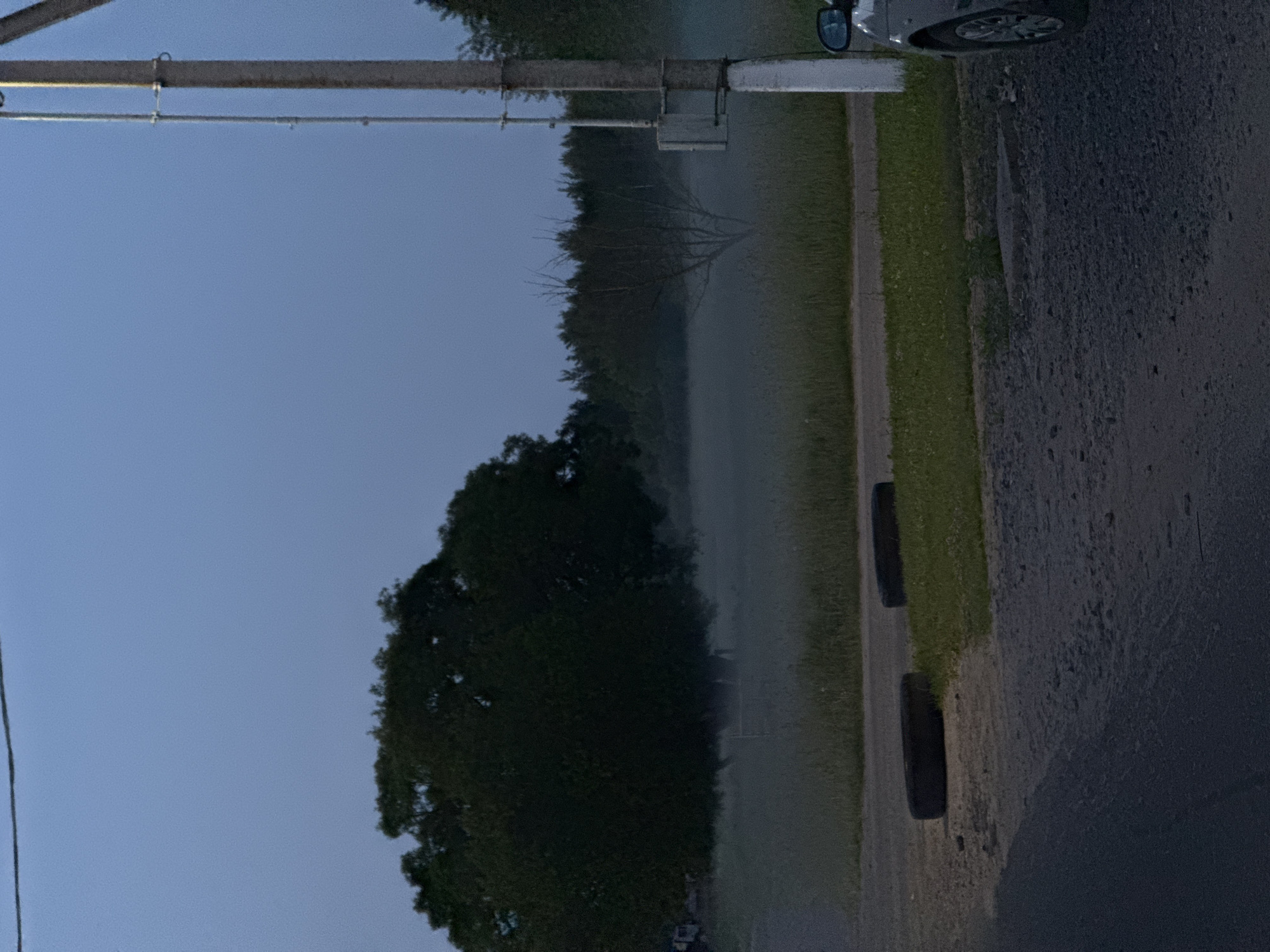 The image, correctly oriented, depicts a quiet rural scene at either dawn or dusk, characterized by soft, diffused lighting. In the foreground on the right, a paved or gravel road is partially visible, with the front wheel and side mirror of a car at the very top right edge, indicating a parked vehicle. Adjacent to the road is a strip of green grass, where two dark, round objects, possibly old tires, rest.
Stretching across the top of the frame is a large, metallic-looking horizontal beam or pipe, secured by a vertical post on the left and featuring various brackets and a small box-like fixture. A thinner line, possibly a wire or pipe, runs parallel beneath it. Farther left, a power line extends diagonally downwards.
The midground is dominated by a dense treeline, which forms a dark silhouette against a pervasive layer of mist or fog that blankets the area beyond the trees. Within this hazy expanse, the stark, bare branches of a leafless tree are visible. The sky above is a muted blue-gray, indicating either early morning, late evening, or an overcast day, with the mist reinforcing the former. No human or animal activity is observed. The scene evokes a tranquil and somewhat atmospheric mood in a rural setting, consistent with the location context of Лабжанскі сельскі Савет, Belarus. No visible text is present.