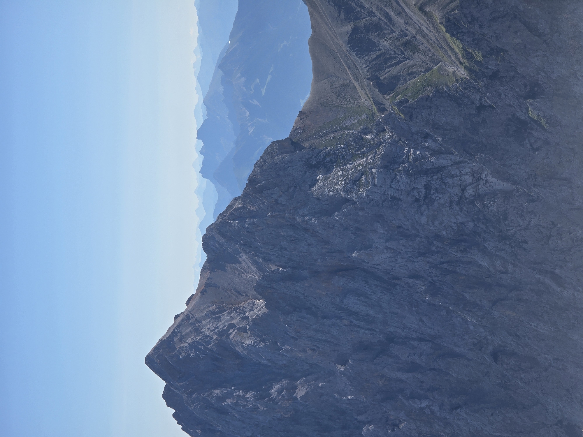 The image depicts a panoramic view of the Alps, with layers of blue mountains receding into the distance under a clear, light blue sky. In the foreground, a large, rugged, grey mountain dominates the left side of the frame. Its steep, rocky slopes are textured with visible crevices and variations in shade, suggesting a rough and uneven surface. The peak of this mountain is sharp and pointed.
To the right of the main mountain, the terrain slopes downwards, revealing a ridge with a lighter, brown and green-colored surface. This ridge is characterized by gentler slopes and scattered patches of vegetation. Further in the background, numerous mountain ranges fade in color and detail, creating a sense of depth and vastness. The atmospheric perspective contributes to the haziness of the distant peaks. There are no people, animals, or man-made structures visible in the scene. The environment is entirely natural and mountainous. The image appears to be taken from a high vantage point, looking out over the mountain landscape. While the exact location within Garmisch-Partenkirchen is not specified by the visual cues, the scenery is consistent with the mountainous terrain found in and around the Bavarian Alps.
