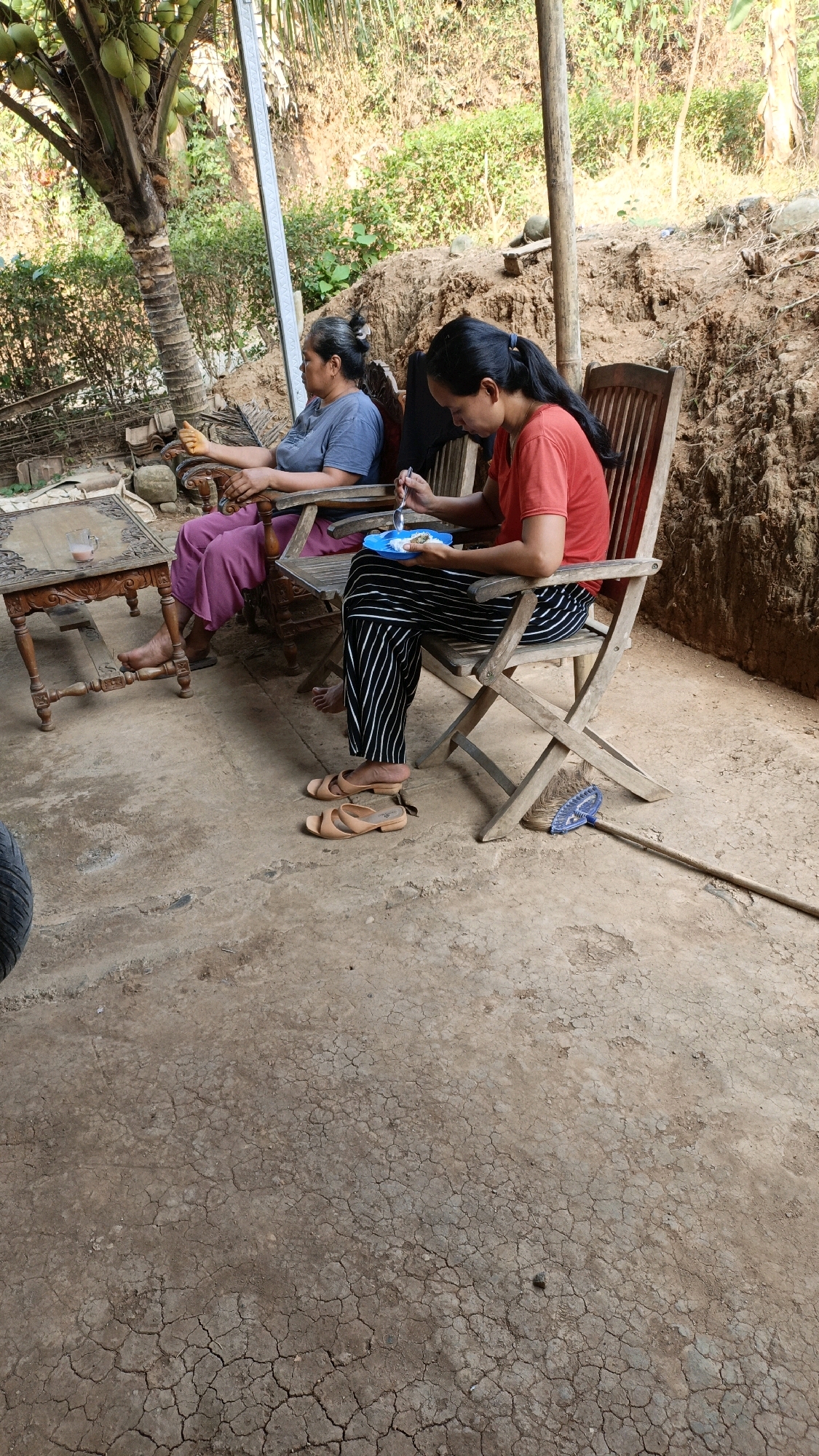 In Suwawal Timur, Indonesia, two women are seated in chairs outdoors, in front of a pile of dirt and greenery. The woman on the left, wearing a blue t-shirt and pink pants, is looking to her left. The woman on the right, in a red t-shirt and black and white striped pants, is eating from a blue plate with a fork. A small table with a glass of drink is next to the woman on the left. A broom with a blue handle lies on the ground near the woman on the right. The ground is cracked and dry.