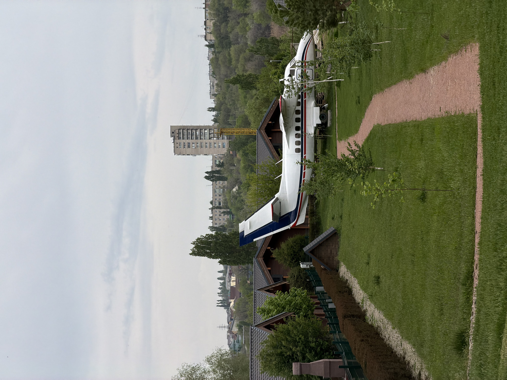 The image depicts a grounded, partially disassembled passenger plane resting on a green lawn among wooden structures, possibly cabins, in what appears to be a park or recreational area in the city of Kamyshin, Russia. A building under construction with a yellow crane is visible in the background. A gravel path runs alongside the airplane. The sky is overcast.