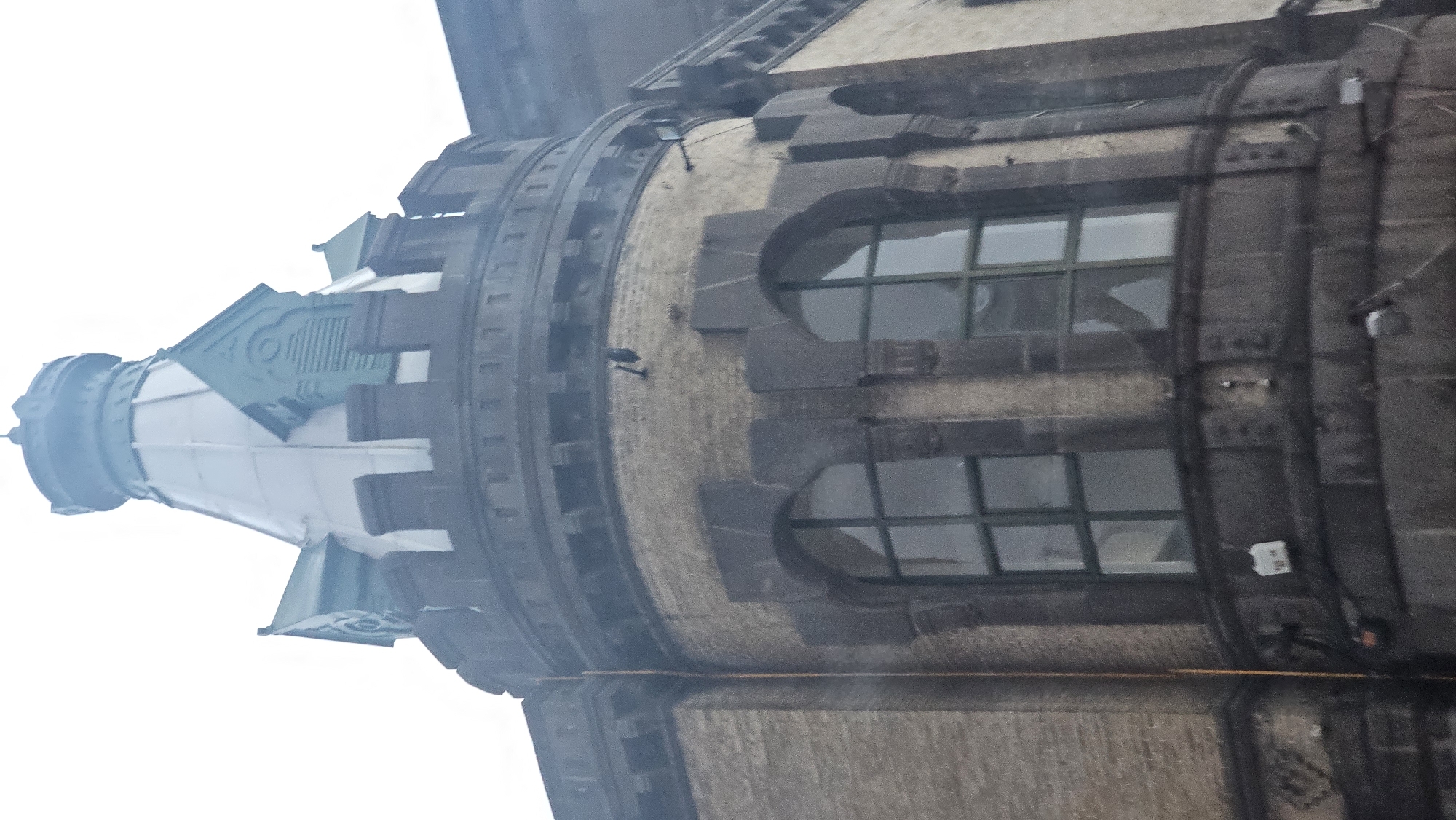 The image captures an upward view of the upper section of a tall, cylindrical stone building against a bright, uniformly white, overcast sky. The building's main body is constructed of light-colored stone or brick, featuring darker, intricately carved stone elements, including ornate window frames and cornices. Several large, rectangular windows with gridded panes are visible. A small, dark bird is perched on a decorative ledge midway up the cylindrical section. The building culminates in a distinctive, multi-tiered conical spire or cupola, which features prominent green-colored accents and intricate detailing, tapering to a small finial at its apex. Wires or cables are visible running down the darker sections of the building's facade. The architectural style suggests a historical urban building. The diffused lighting indicates an overcast day. No human activity or visible text is present. Given the context, this structure is located in Puebla, Mexico.