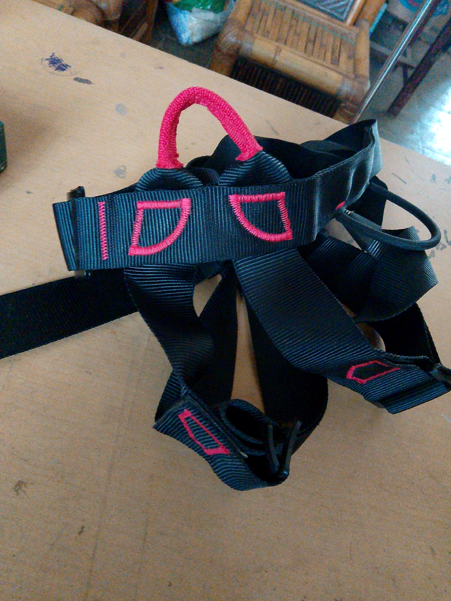 The image shows a black climbing harness with pink stitching and a pink loop, lying on a light brown surface. The harness has the letters "IDD" embroidered in pink on a black strap. The surface appears to be a table or countertop, with some smudges and what looks like dried paint or ink stains. In the background, out of focus, is a wooden chair and some blurred items, possibly bags. The scene appears to be indoors, in a room that might be a workshop or storage area. There is no indication of the specific location in Kuripan Utara, Indonesia, as the image does not contain any recognizable landmarks or contextual clues.