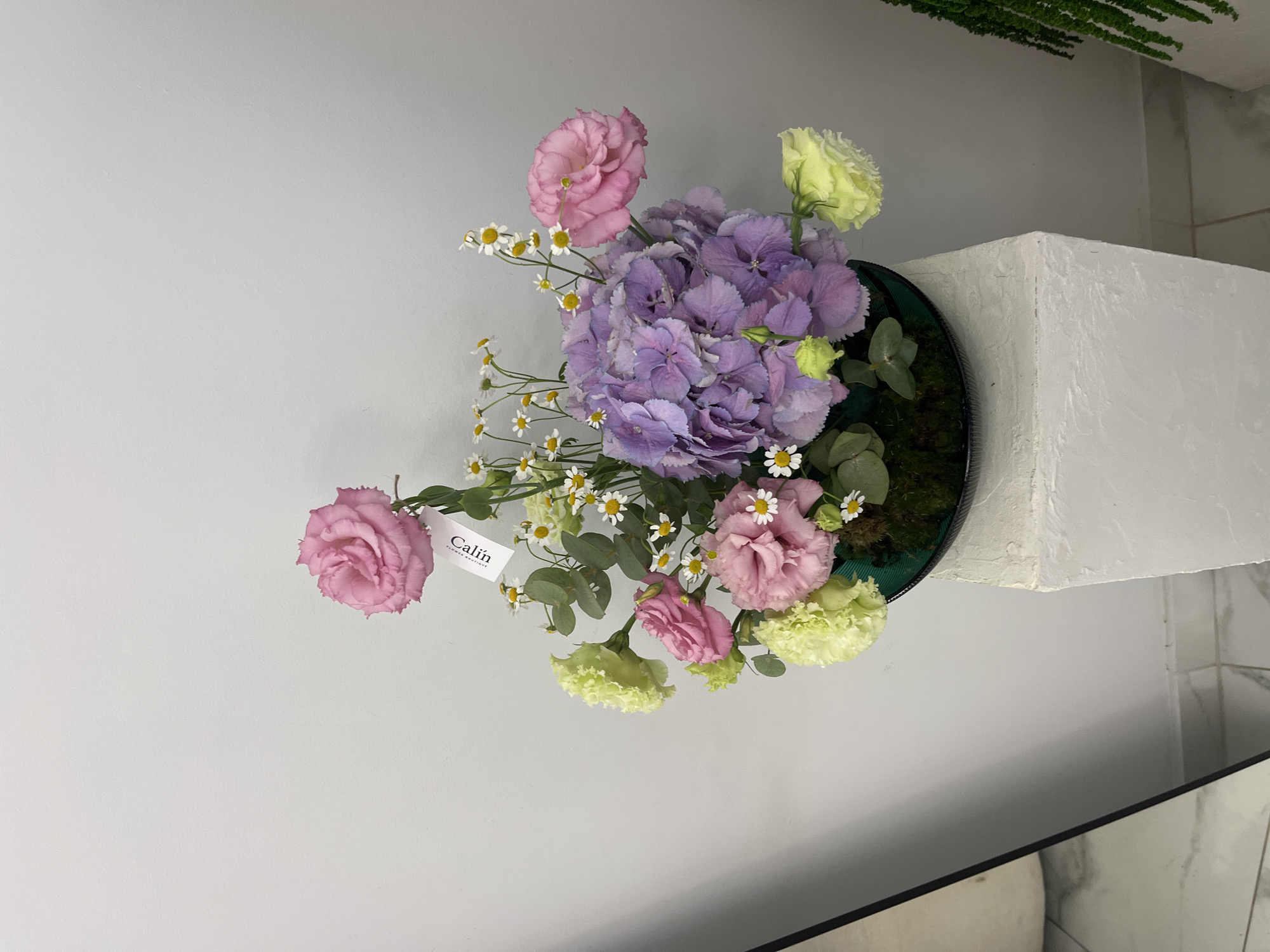 In Kaspiysk, Russia, a floral arrangement sits on a textured white pedestal. The arrangement features a large purple hydrangea as its centerpiece, surrounded by pink roses, pale yellow carnations, and delicate white daisies. A small tag labeled "Calin" is attached to one of the pink roses. The background is a plain white wall. To the left, a portion of a reflective surface, possibly a mirror or glass, is visible, showing a marble-tiled floor. To the right, a green fern plant is partially visible.