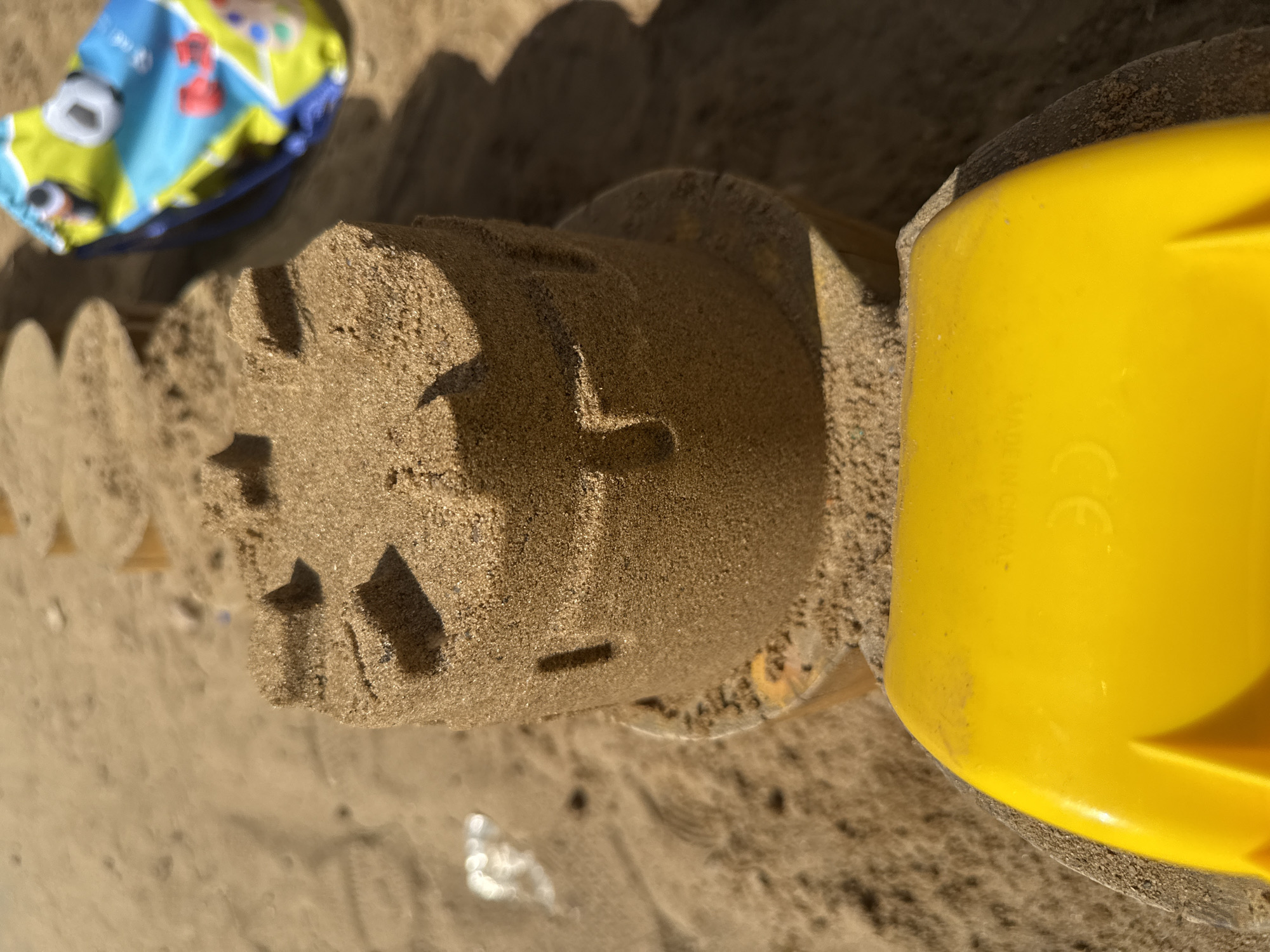 The image shows a sandcastle, likely made on a beach. The sandcastle is cylindrical with patterned sides, bearing impressions of crosses and squares. A yellow plastic bucket is nearby, partially obscuring the sandcastle. In the background, there is a blue and yellow patterned beach toy. The environment appears to be sandy. There is no indication that the image was taken in Lyubertsy, Russia.