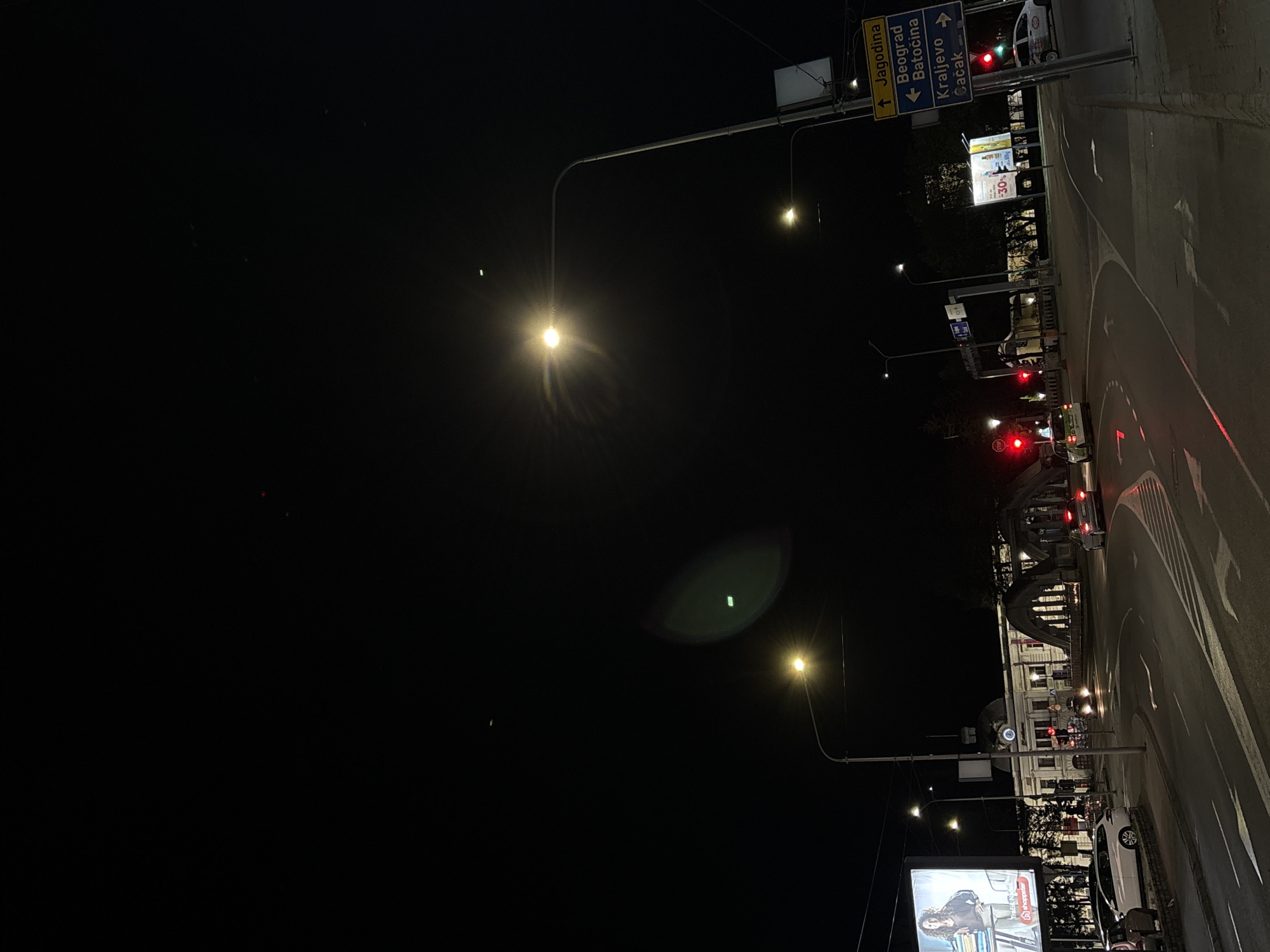 The image depicts a night scene in Kragujevac, Serbia. The location is near the main train station. A multi-lane road curves around a traffic island. Streetlights illuminate the scene. Traffic lights are visible, some displaying red. A directional sign indicates directions to Jagodina, Beograd (Belgrade), Batočina, Kraljevo, and Čačak. Several cars are present. A billboard is on the right side of the image. The main train station building is visible in the background.