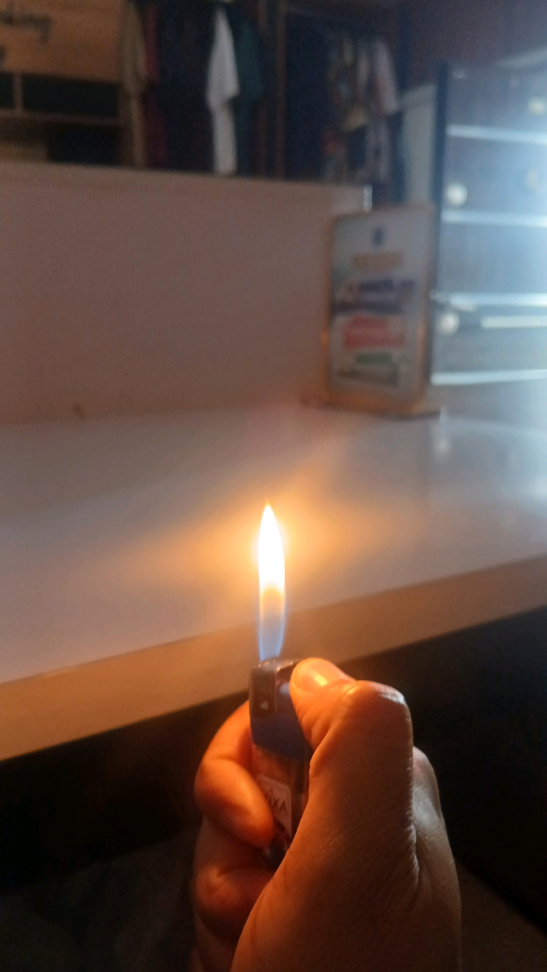 A close-up shot shows a hand holding a lit lighter. The flame from the lighter is bright orange and white, illuminating the hand and the surrounding surface. The lighter itself is blue and appears to have some red markings on it. The background is blurred, but suggests an indoor setting with shelves and possibly some clothing items. The overall lighting is dim, with the lighter's flame being the primary light source. The location in the city of Gura Top, Nigeria cannot be determined from the image alone.