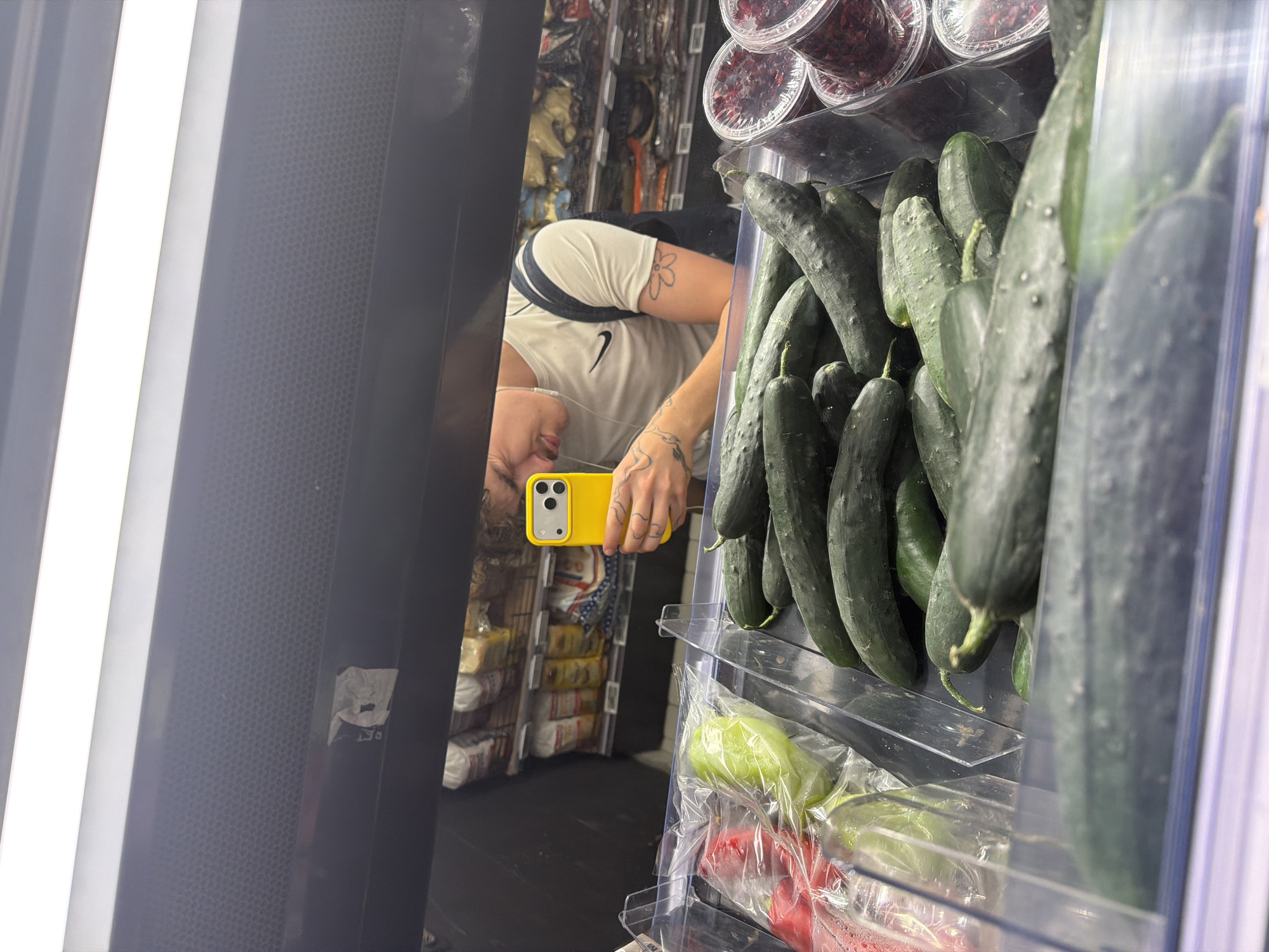 A person is reflected in a refrigerated display case in what appears to be a store, possibly located in Vracar Urban Municipality, Serbia. The person is holding a yellow smartphone and is likely taking a photo of the produce within the case. The shelves contain cucumbers and packaged vegetables. Above the cucumbers, there are containers with what look like dried cranberries. The reflection shows grocery items in the background. The left side of the frame includes part of the display case, with its built-in lighting.