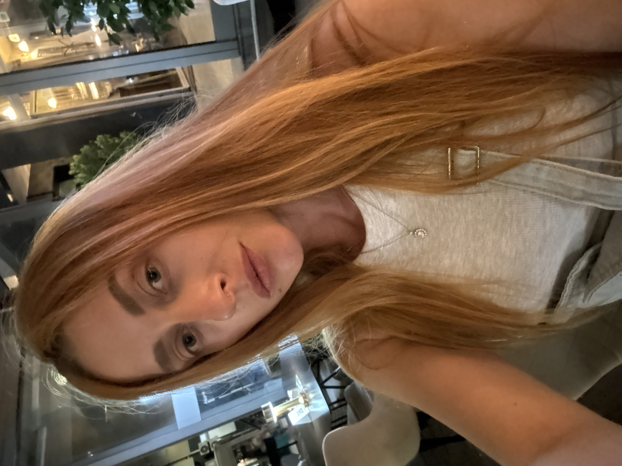 A woman with long, auburn hair is taking a selfie in an indoor setting that appears to be a restaurant or cafe. She is wearing a light-colored tank top with suspenders and a small silver necklace with a pendant. The background shows tables, chairs, and large windows with views of the outdoors, suggesting a modern establishment. The lighting is dim, characteristic of an evening setting. The location is the city of Budva, Montenegro.