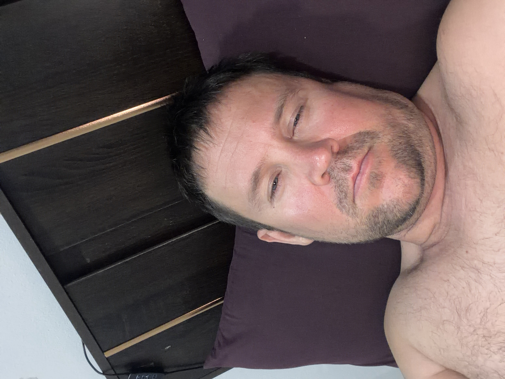 A man lies on a dark purple pillow, resting his head on a dark brown headboard. His eyes are partially closed, and his mouth is slightly open. He has stubble on his chin and a light beard. His chest and shoulders are visible, and they are covered in dark hair. The headboard has a thin, golden stripe running down the middle. The background is a plain light blue wall. There is a black cable with a small black object attached to it running along the top of the headboard, near the left edge of the image. The scene appears to be indoors, likely in a bedroom.