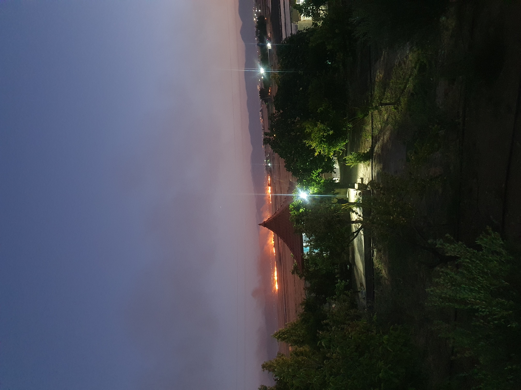 The image shows a dusk scene in Kheyrabad, Iran. In the foreground, there are several trees, some of which are illuminated by artificial lights. A pathway leads into the scene, with more trees and vegetation on either side. In the middle ground, there is a building with a conical roof, also illuminated by lights. Beyond the building, a large area is visible, with a distant fire or multiple fires burning, creating a hazy glow in the sky. In the background, there are mountains and a faint line of lights, suggesting a settlement or town. The sky is a gradient of deep blue and purple, with a few faint clouds.