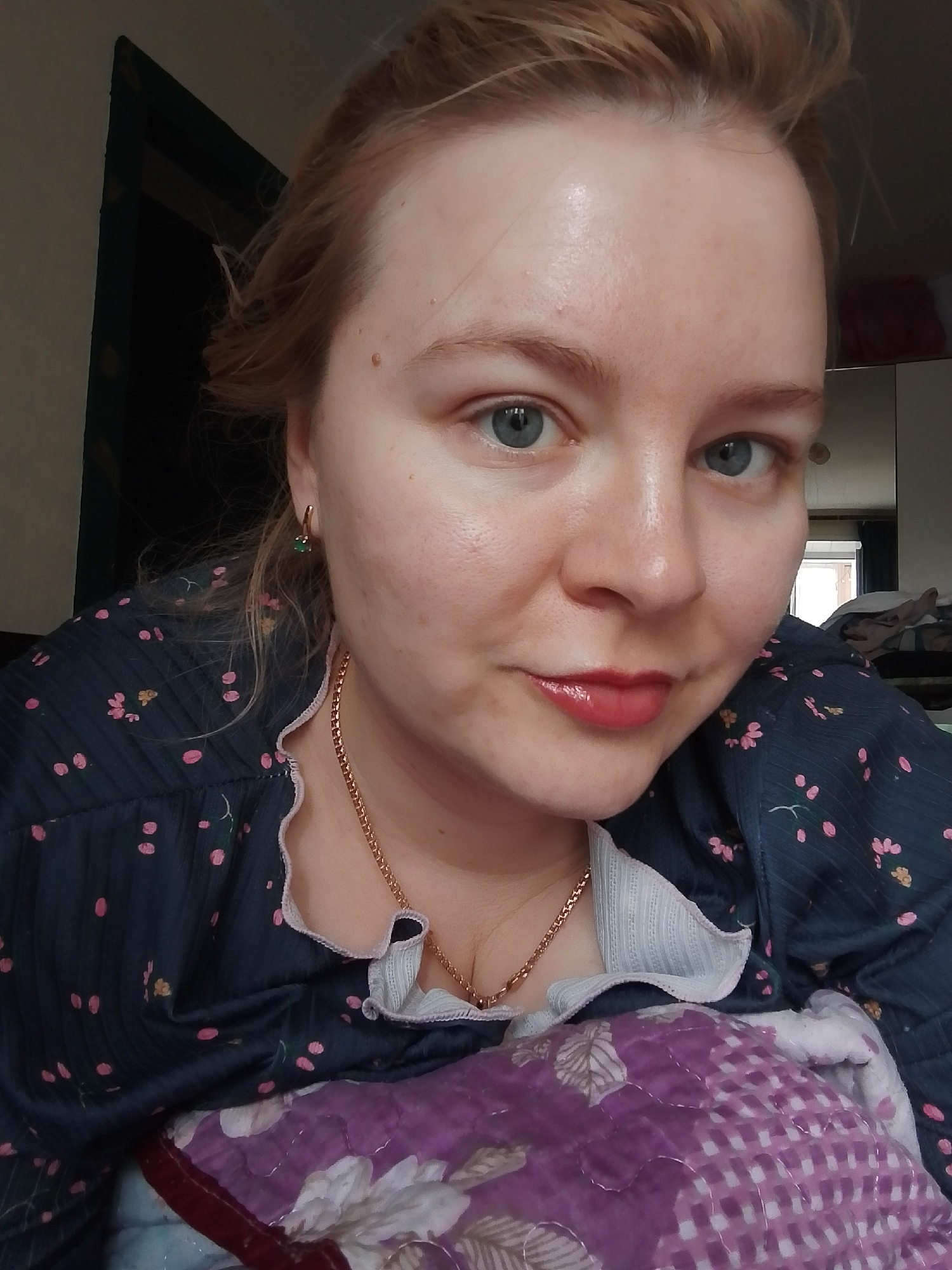 A medium close-up shot depicts a fair-skinned woman with blue eyes and light red hair. She is wearing a navy blue shirt with a small, floral pink pattern and a gold necklace. She appears to be lying down, as a purple and white floral patterned blanket is visible in the foreground. A small, green earring is seen on her left ear. The background is blurry, but a dark doorway is visible on the left and indistinct objects are visible on the right. There is no clear indication in the image that it was taken in Izhevsk, Russia, or any other specific location.