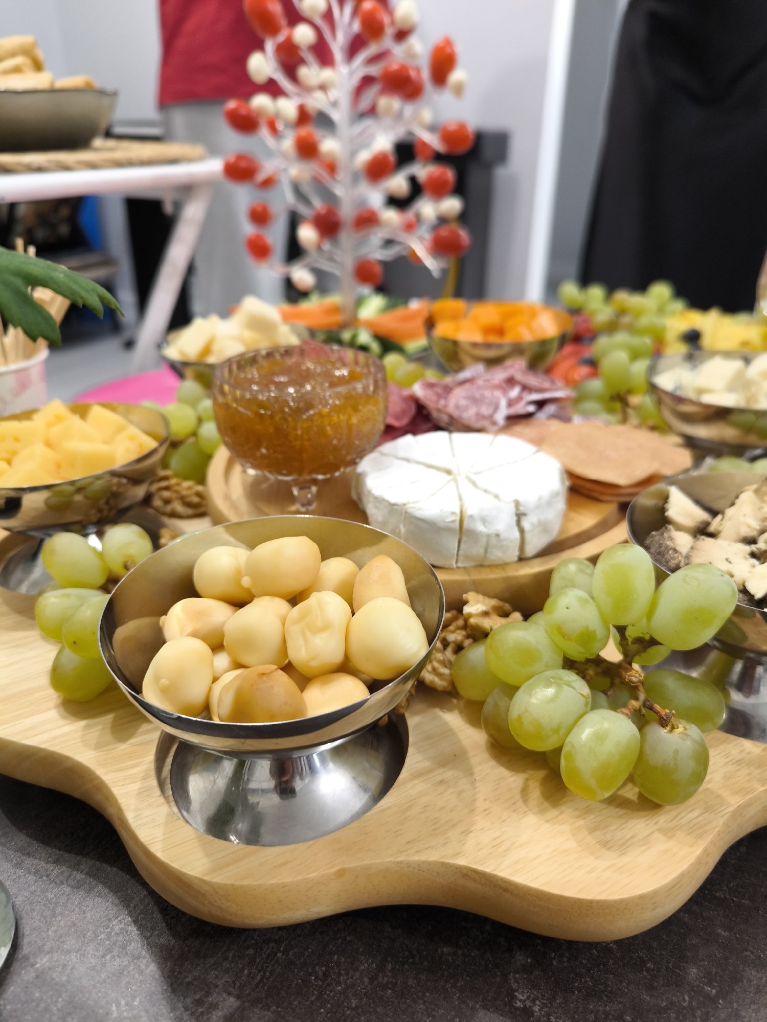 A charcuterie board on a dark gray surface features various items, including a bowl of pale, round objects (likely cheese balls or macadamia nuts), green grapes, Brie cheese, jam in a footed glass container, and various sliced meats and cheeses. The items are arranged on a light-colored wooden board with silver bowls and accents. In the background, there is a decorative tree-like structure with red and white spheres, and blurred figures of people are visible. The location is Fryazino, Russia.