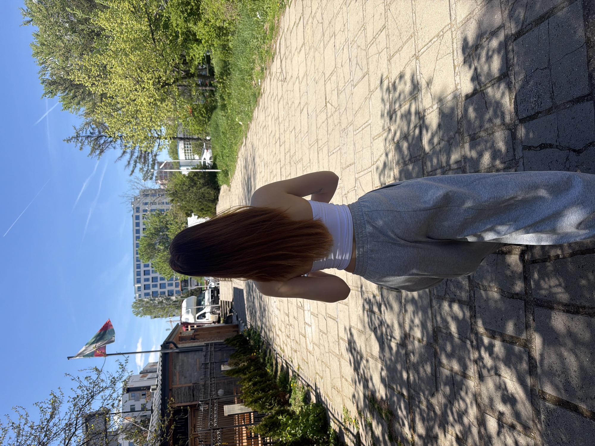 A medium shot depicts a person from behind, standing on a stone-paved path flanked by foliage and trees. The person is wearing a white top and gray sweatpants. The path is shaded by trees, casting shadows on the pavement.
In the background, buildings are visible, including a modern high-rise and a row of smaller, older buildings. A flag is displayed on a pole.
Based on the flag and architectural style, this scene is likely located in Sofia, Bulgaria. Specifically, the flag appears to be the national flag of Bulgaria, and the architecture is consistent with that found in various parts of the city.