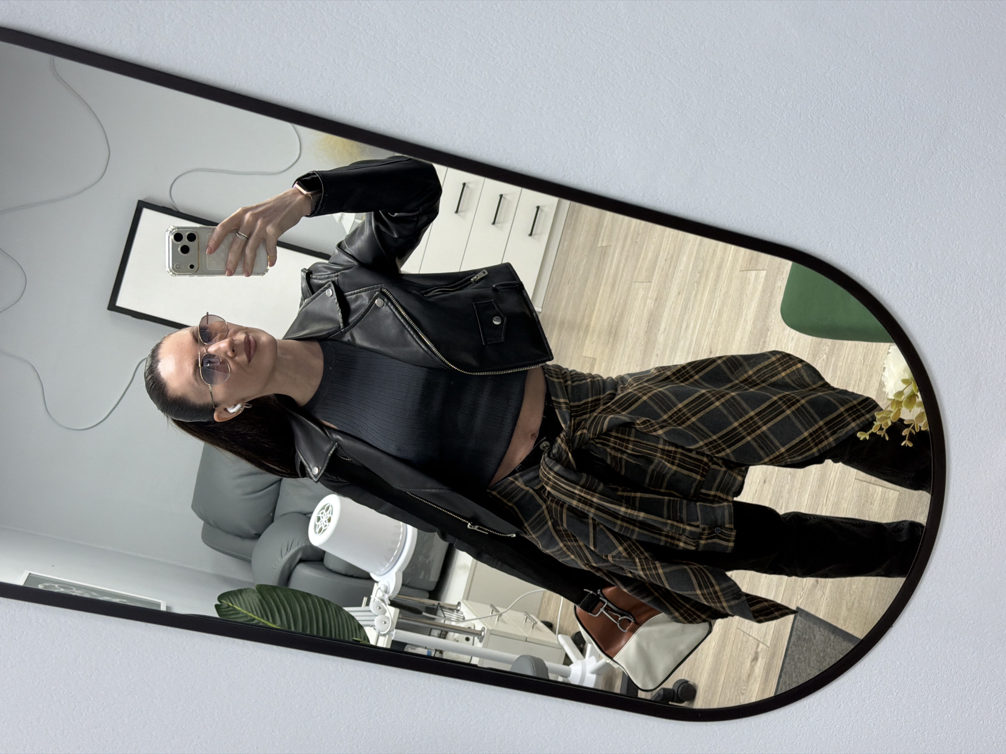 A woman is taking a selfie in a mirror. She is wearing sunglasses, a black leather jacket over a gray top, and black pants. A plaid shirt is tied around her waist. She is holding a phone in her right hand. In the background, there is a gray chair, a white cabinet with drawers, a framed picture on the wall, a floor lamp, and a plant. The location appears to be an interior space, potentially in Perm, Russia.