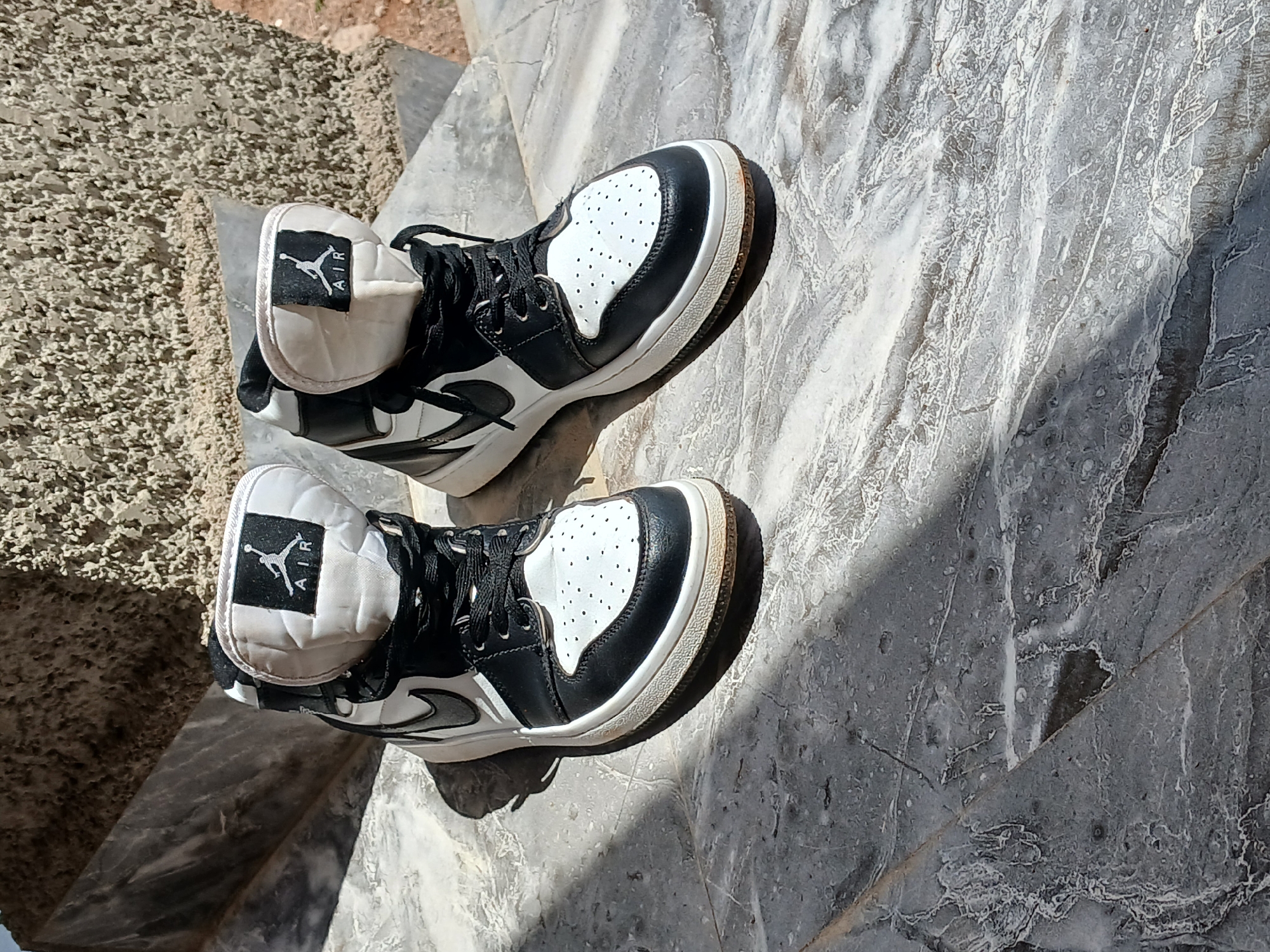 A pair of black and white Air Jordan 1 high-top sneakers are positioned on a marble-like surface. The sneakers are slightly dirty. The background consists of a rough, textured wall with brown and gray tones, and a diagonal shadow from an unseen object. The scene is illuminated by bright sunlight, creating distinct shadows on the surface. The location is Katon Rikkos, Nigeria.