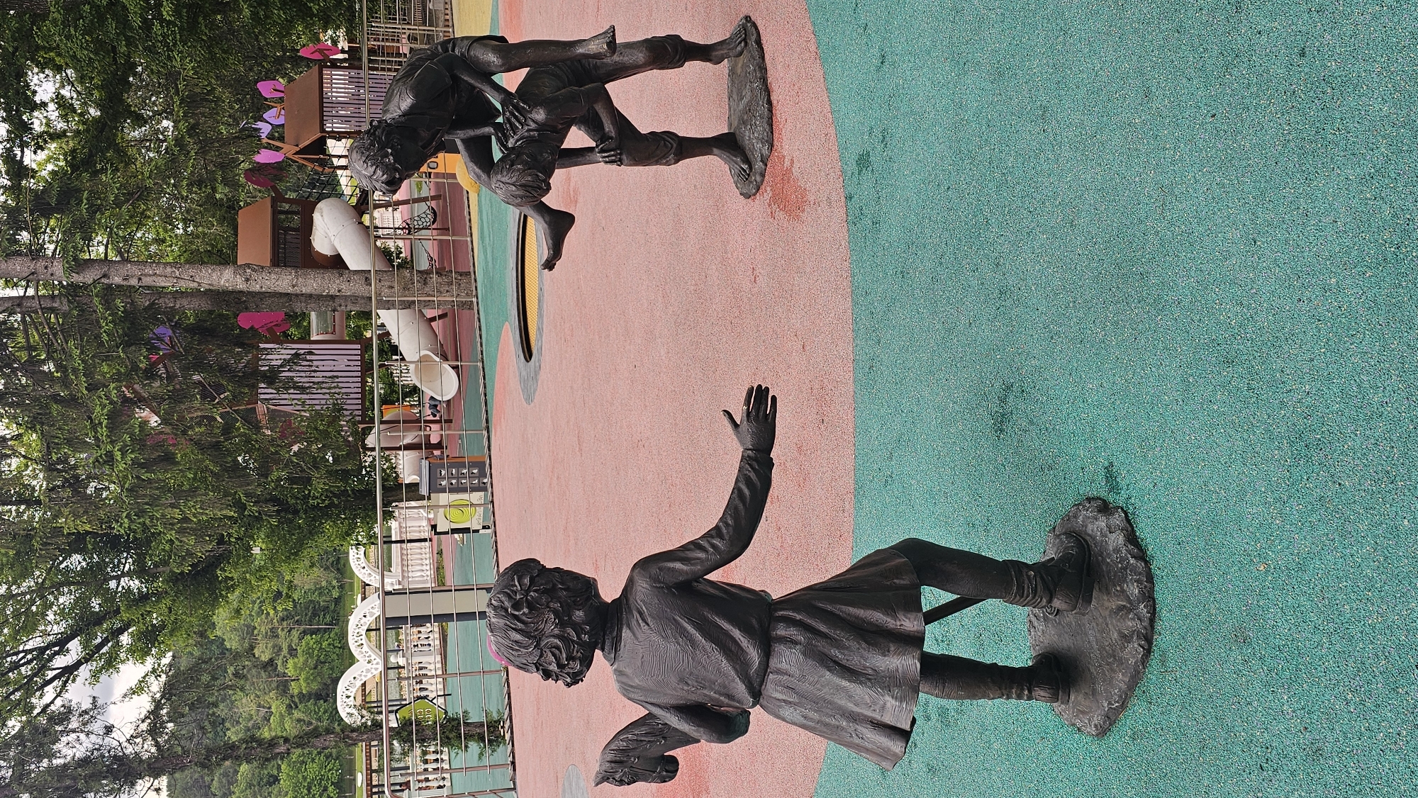 The image depicts a public playground located in Alikonovka, Russia. The playground features several bronze sculptures of children. In the foreground, a sculpture of a young girl, seen from the back, is holding a toy horse. Her right arm is extended forward, with her palm open. To the right, two other sculptures depict children engaged in play. One child is crouched down, and another is positioned on their back, appearing to be in a playful tussle. The ground of the playground is a soft, textured surface divided into two sections: a predominantly pink area and a turquoise-green area. In the background, elements of a playground structure with slides and colorful accents are visible, along with trees and foliage, suggesting a natural, park-like setting. A body of water and distant hills are visible beyond the playground.