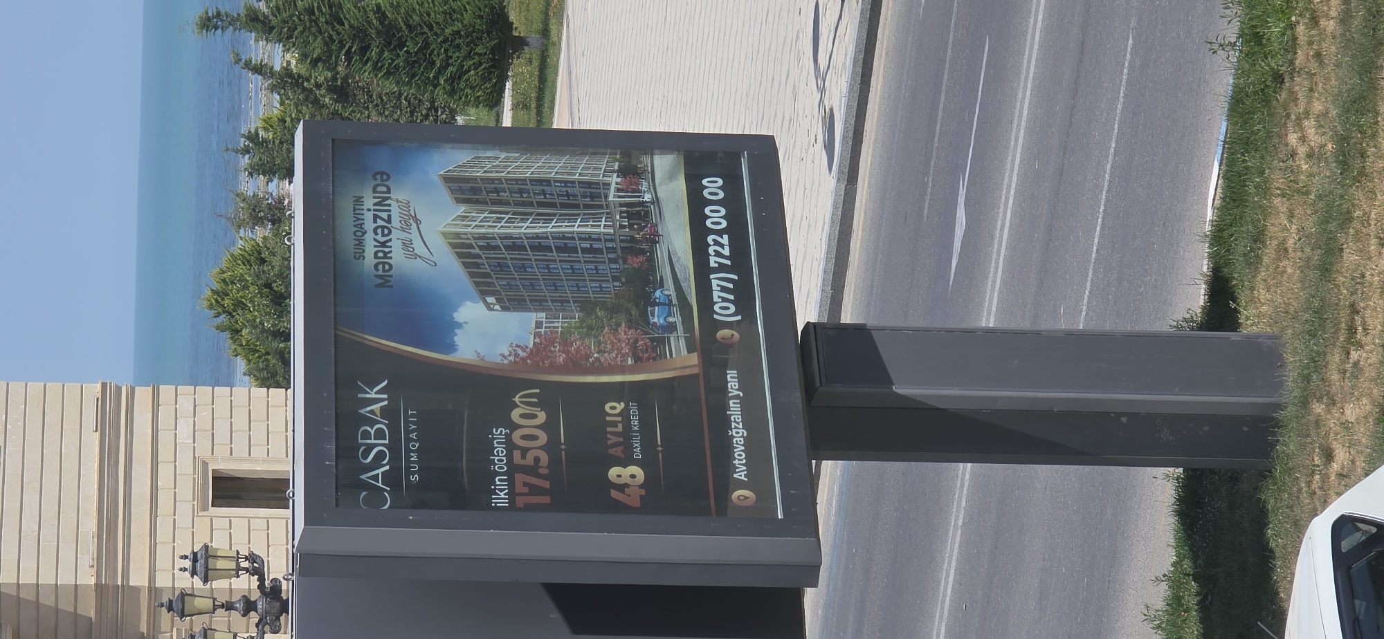 The image shows a billboard advertising real estate in Sumgayit, Azerbaijan. The billboard features a rendering of a modern apartment complex, with text in Azerbaijani indicating "CASBAK SUMQAYIT" as the developer and "SUMQAYITIN MƏRKƏZİNDƏ yeni həyat" which translates to "New life in the center of Sumgayit". Below the building rendering, it shows "ilkin ödəniş 17.500₼" (initial payment 17,500 AZN) and "48 AYLİQ DAXİLİ KREDİT" (48-month internal credit). A phone number, (077) 722 00 00, and a location description "Avtovağzalin yanı" (next to the bus station) are also visible. The billboard is mounted on a tall, dark grey pole and is situated on a roadside with green grass and trees in the background, with the Caspian Sea and a building facade visible in the far distance.