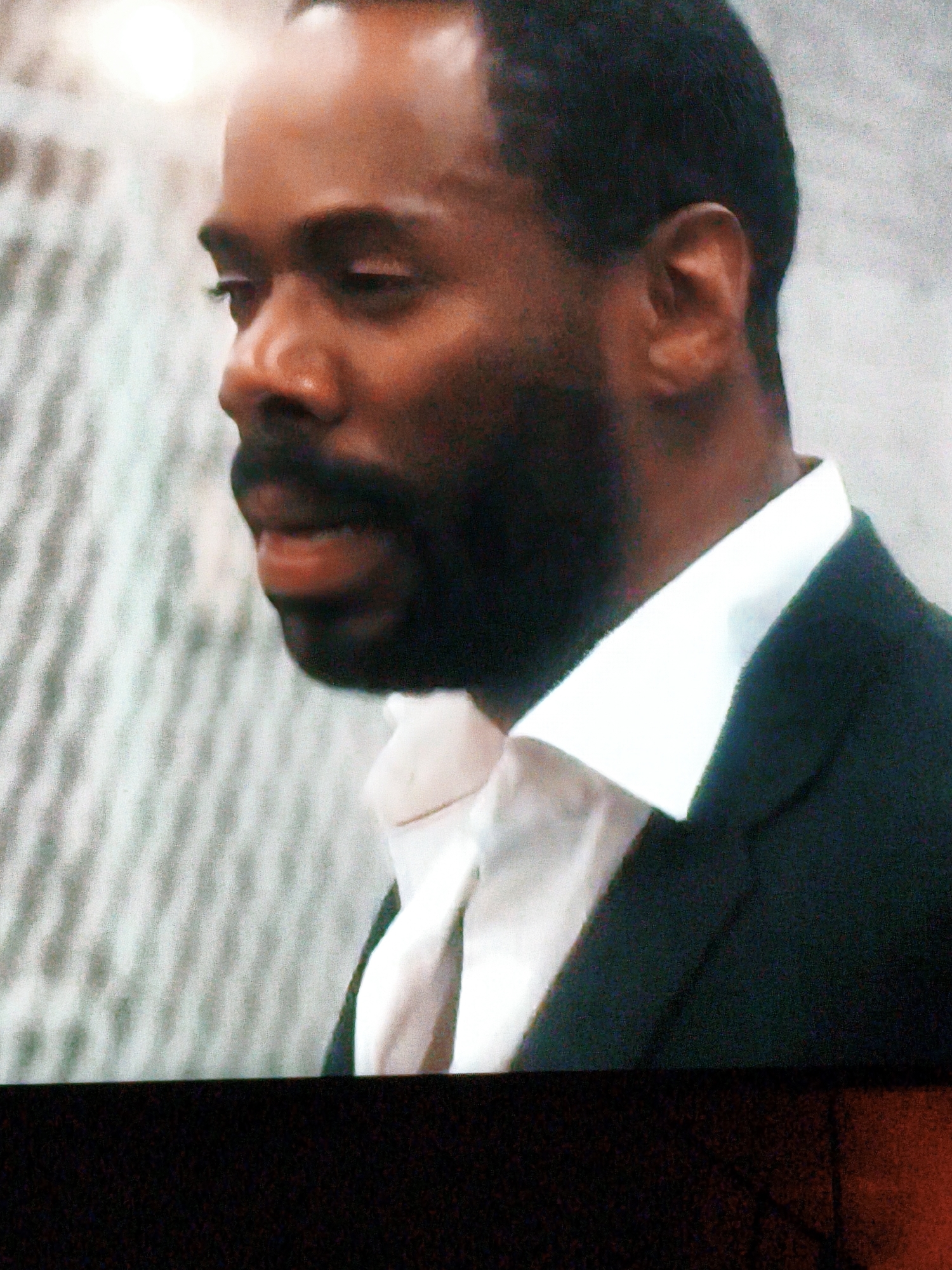 A medium shot shows a Black man in profile, looking to the left. He has dark skin, short dark hair, and a beard. He is wearing a white collared shirt and a dark suit jacket. The background is out of focus and appears to be a wall with a textured pattern. The lighting is bright, creating a highlight on his forehead. There is no indication of the city of Vladivostok, Russia in the image.