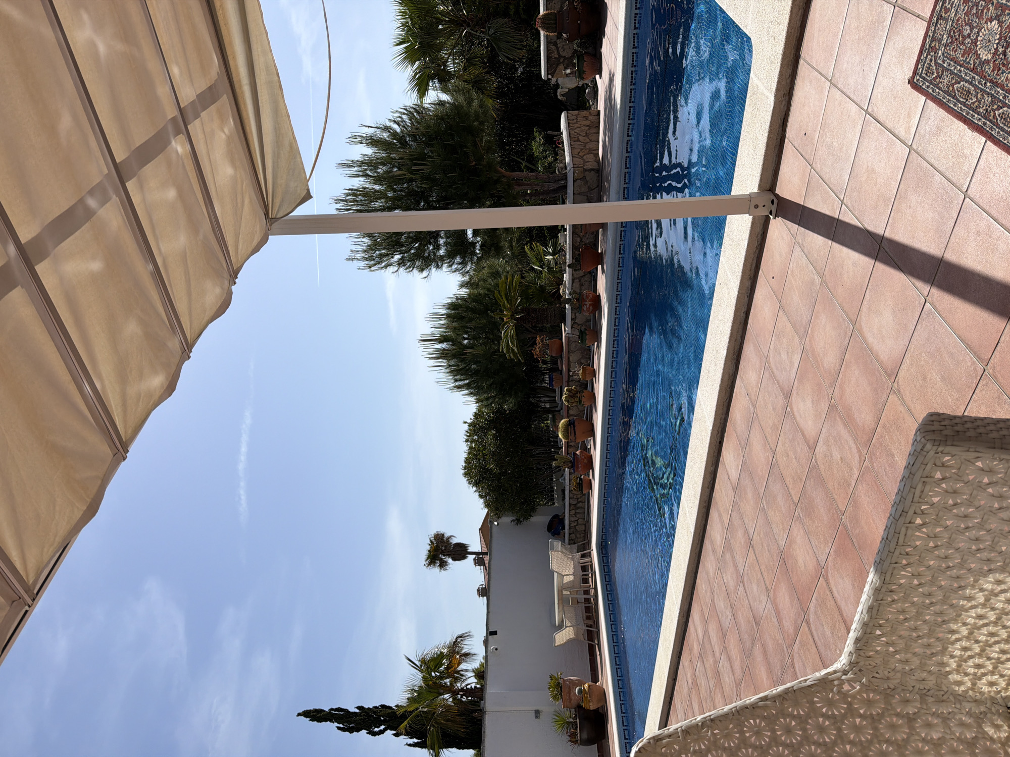 The image depicts an outdoor scene in Mont-roig del Camp, Spain, featuring a swimming pool adjacent to a tiled patio. The pool water is blue, reflecting the sky. A beige awning provides shade over the area. Plants and trees line the perimeter of the pool, and a white wall with additional greenery is visible. Wicker furniture is partially visible in the foreground. A rug sits on the tiled patio, with reddish-brown tiles. The sky is mostly blue with some cloud formations. The angle of the shot is rotated clockwise.