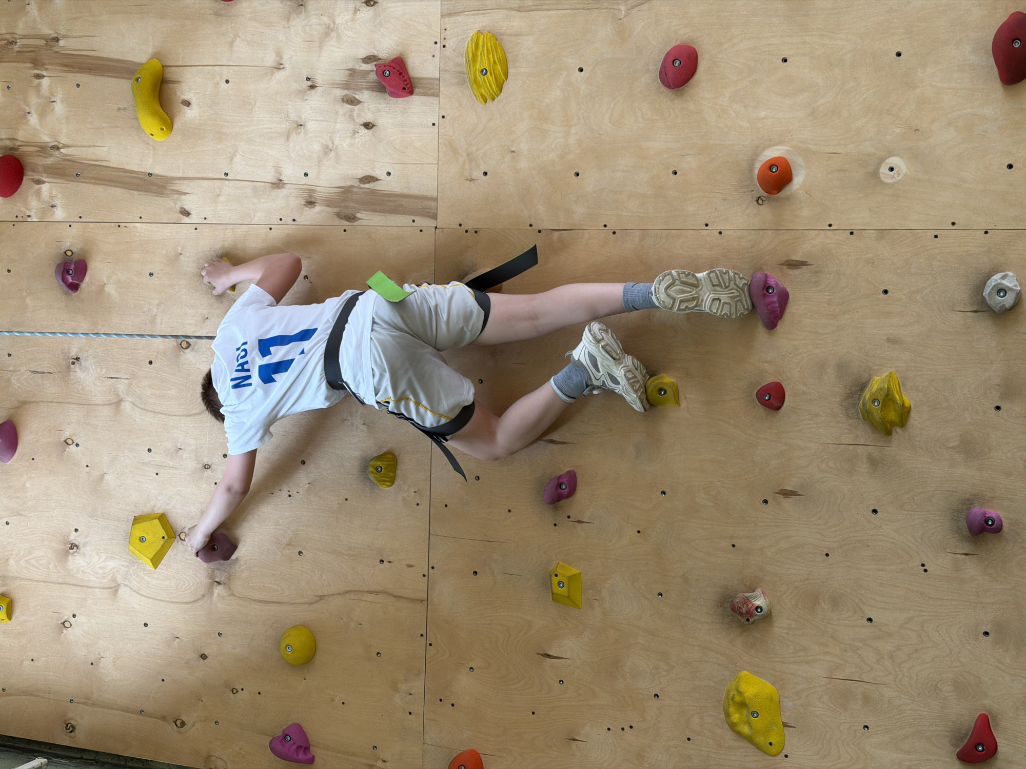 A young person wearing a white t-shirt with the number "11" on the back, grey shorts, and climbing shoes is suspended on a climbing wall. The climbing wall appears to be constructed of plywood and features an array of brightly colored climbing holds of various shapes and sizes. The person is secured with a climbing harness. The location of this scene is not explicitly mentioned. Therefore, I cannot determine if it's in Izhevsk, Russia.