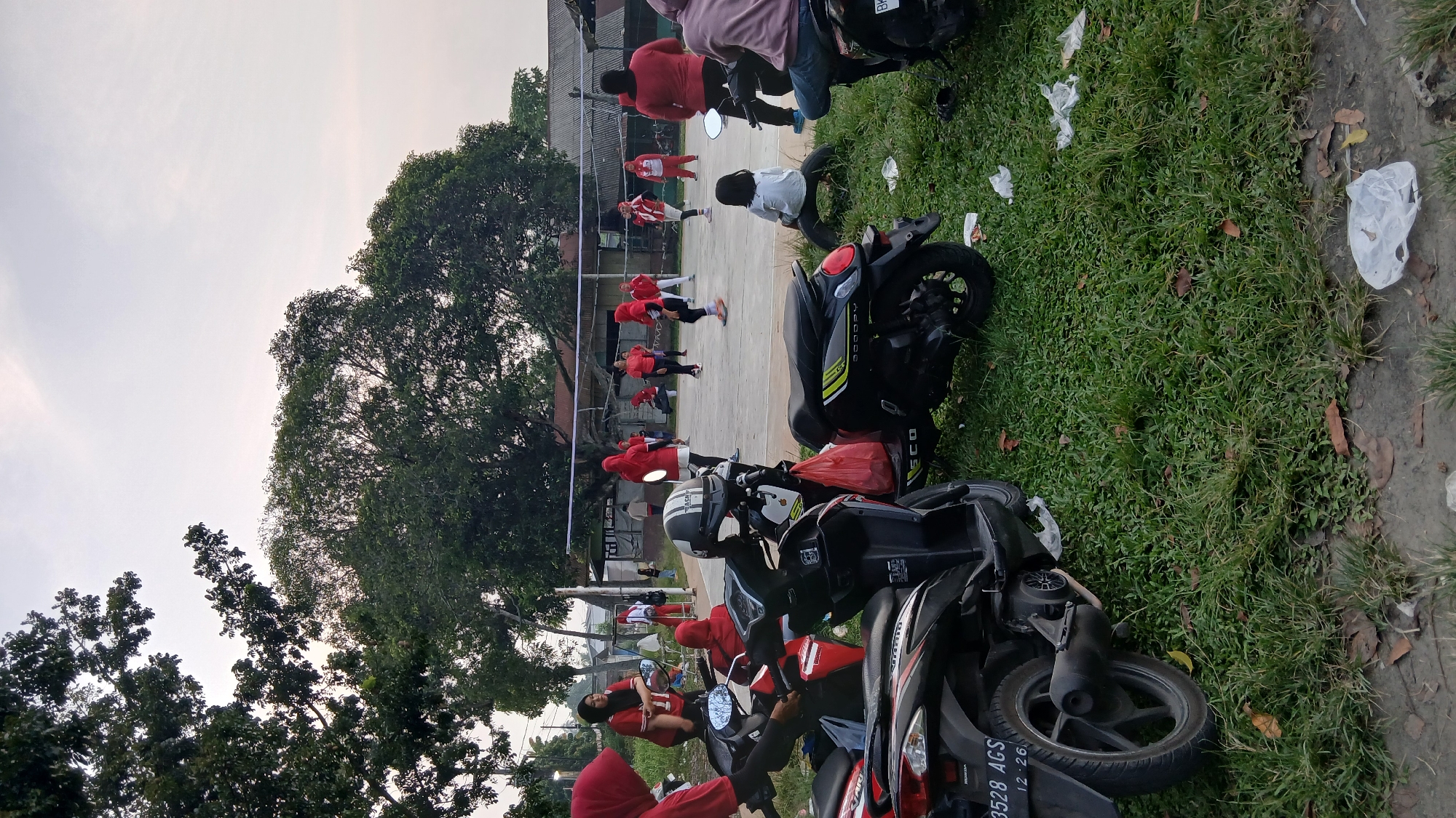 In Klumpang Kebun, Indonesia, a group of women are playing volleyball on an outdoor court. The scene is captured in the late afternoon, with the sky overcast and trees providing shade around the perimeter of the court. Several motorcycles are parked in the foreground, with one black scooter bearing the "SCOOPY" logo. The court itself is a paved surface, with a volleyball net stretched across the middle. The players, all wearing red outfits, are actively engaged in the game. Some are positioned near the net, while others are spread out on the court. In the background, there are simple buildings and more trees. The overall atmosphere suggests a community gathering or recreational activity taking place in a park-like setting.