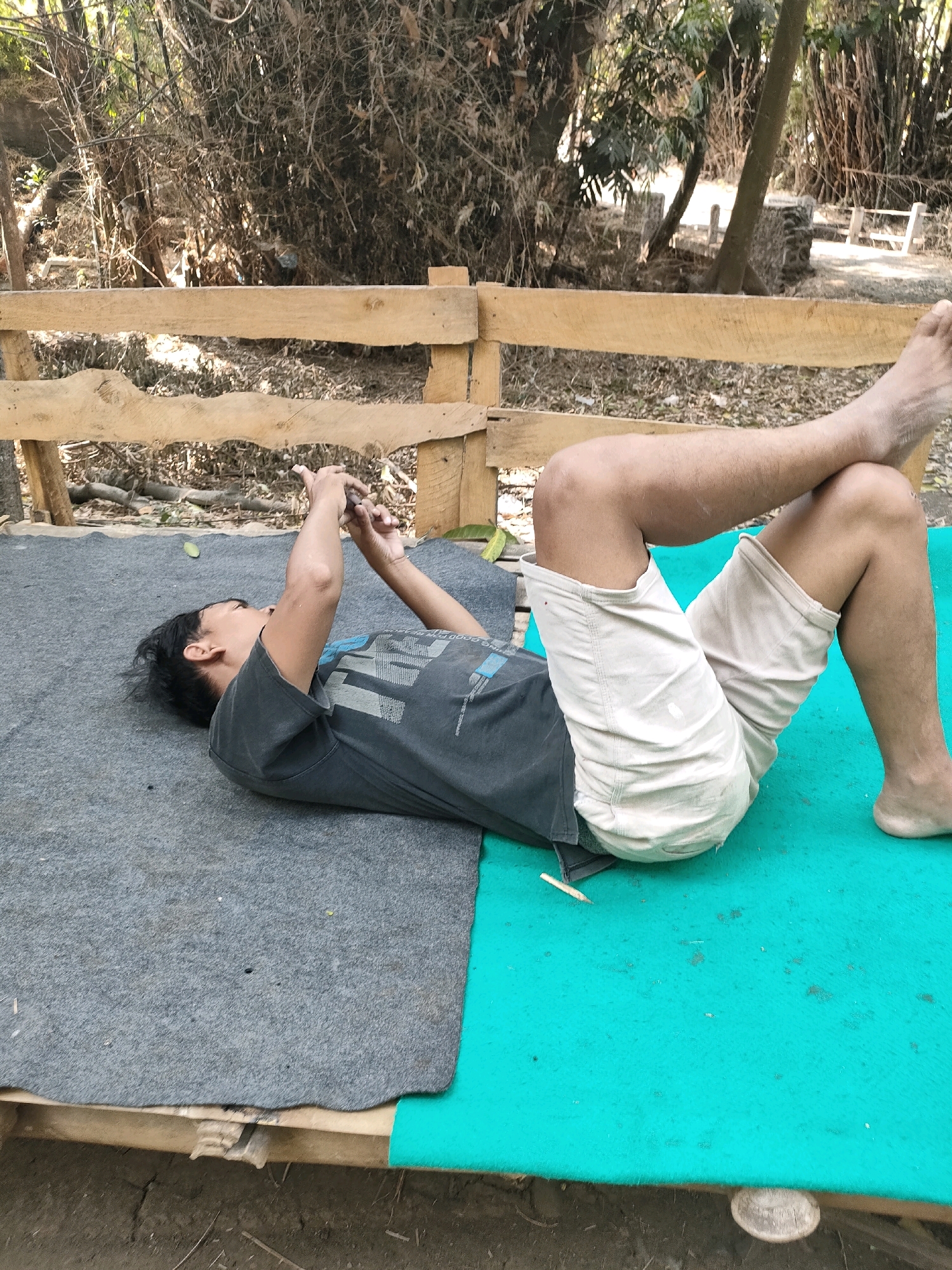 A young man with black hair is lying on his back on a grey mat. He is wearing a dark grey t-shirt with a graphic print and light-colored shorts. His eyes are focused on a smartphone held in his hands, which are positioned above his face. The mat is situated on a wooden platform, partially covered by a turquoise mat on the right side. In the background, a wooden fence runs horizontally. Beyond the fence, there are trees and dense foliage, suggesting an outdoor, possibly rural or natural setting, consistent with the area of Tongas Kulon, Indonesia. The lighting indicates it is daytime.