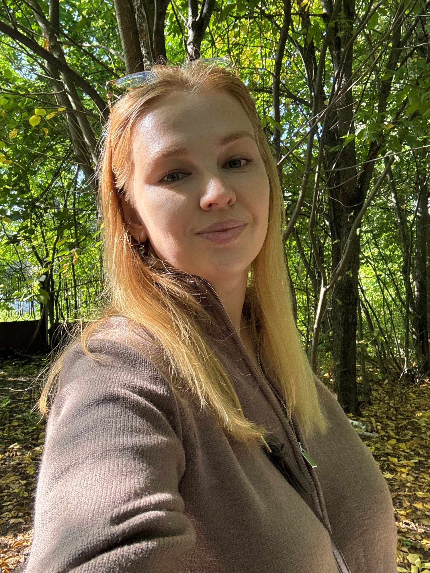 The image is a close-up, front-facing selfie of a fair-skinned woman with shoulder-length blonde hair and light-colored eyes, taken outdoors in a wooded area. She is wearing a brown zip-up sweater. Sunglasses are perched on top of her head. The background consists of trees with green and yellow leaves, suggesting an autumn season. The ground is covered with fallen leaves. The lighting indicates it is daytime, with sunlight filtering through the trees. The location is described as the city of Yaroslavl, Russia, specifically within a park or wooded natural area.