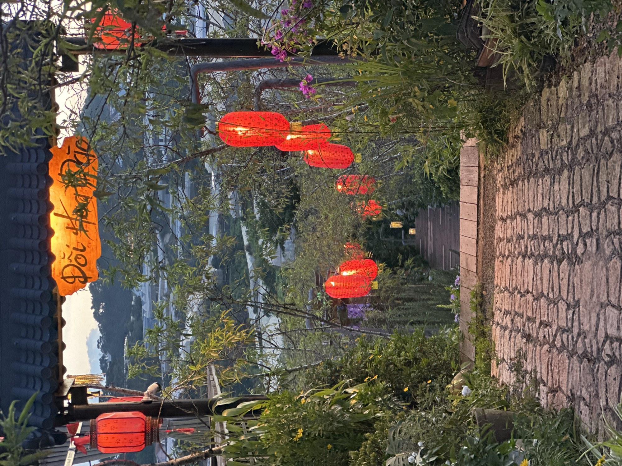 The image depicts a pathway lined with lush greenery and adorned with numerous red paper lanterns, creating a festive and inviting atmosphere. The pathway itself is paved with rough-hewn stones, suggesting an outdoor, perhaps rustic, setting. In the background, a valley unfolds with terraced landscapes, possibly rice paddies or tea plantations, hinting at a rural or mountainous region. The scene is illuminated by the warm glow of the lanterns, indicating that it is either dusk or evening. The lighting from the lanterns casts a soft, ambient light, and some are visibly lit from within.
To the left of the image, a traditional-looking roofline with dark tiles is visible, and an illuminated orange paper lantern with handwritten text hangs from it. The text is in an Asian script, likely Vietnamese given the location context of Lộc Quý, Vietnam. The overall mood is serene and tranquil, with a touch of cultural charm. The time of day is suggested by the soft light and the illuminated lanterns, likely sunset or early evening. The vegetation is abundant, with trees and bushes framing the pathway and lanterns. There are no people visible in the image. The setting appears to be a scenic outdoor space, possibly a garden, a restaurant, or a pathway leading to a scenic viewpoint.