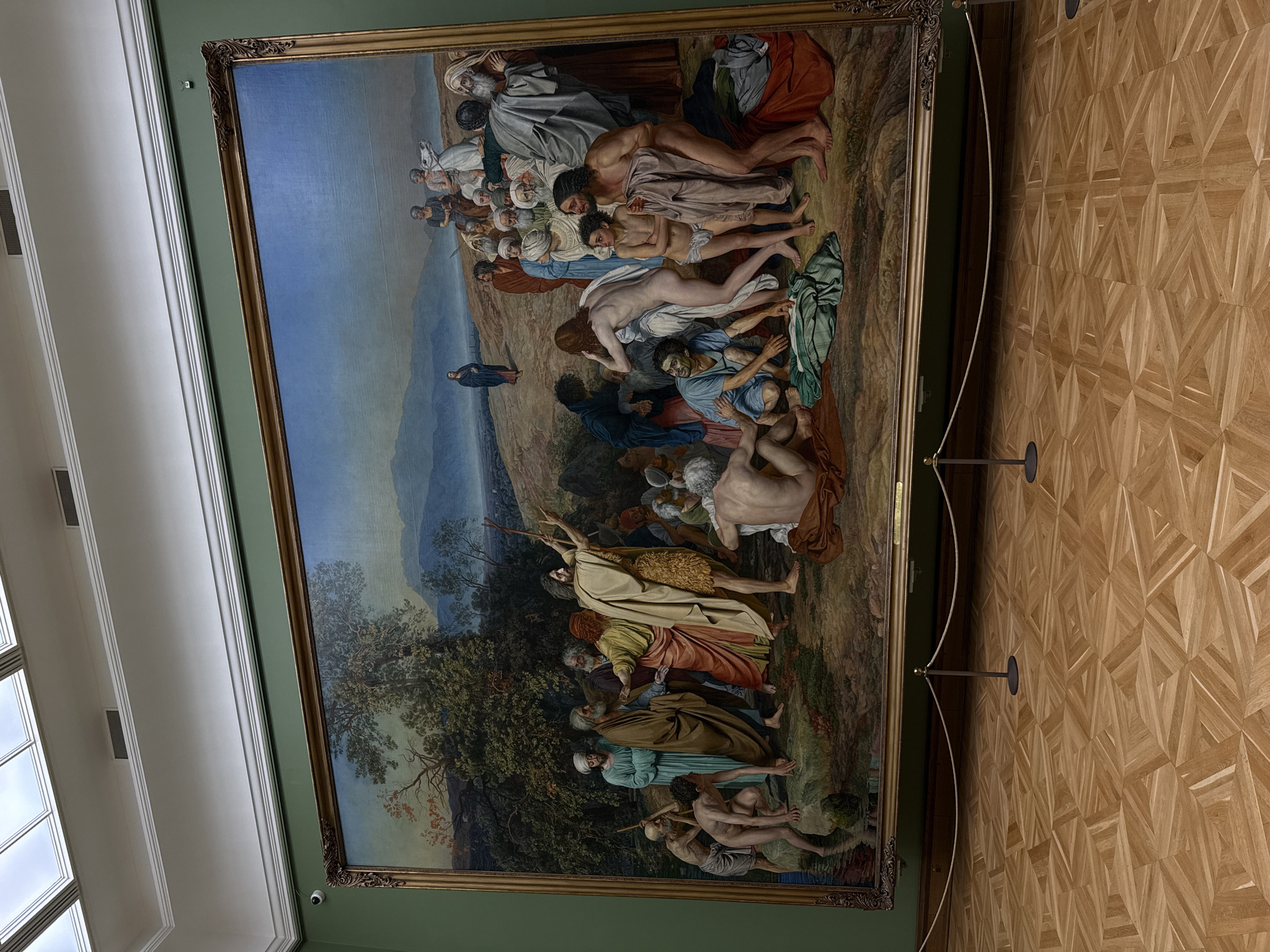 A large, framed painting hangs on a green wall in a gallery setting. The painting depicts a biblical or historical scene with numerous figures in various states of distress and activity, set against a landscape backdrop. The subjects in the artwork are people, some clothed, and some nude. The environment includes trees and mountains. The actions in the painting include people surrounding others and people laying around the landscape. The gallery has a patterned wooden floor. The painting is likely located in the city of Moscow, Russia.