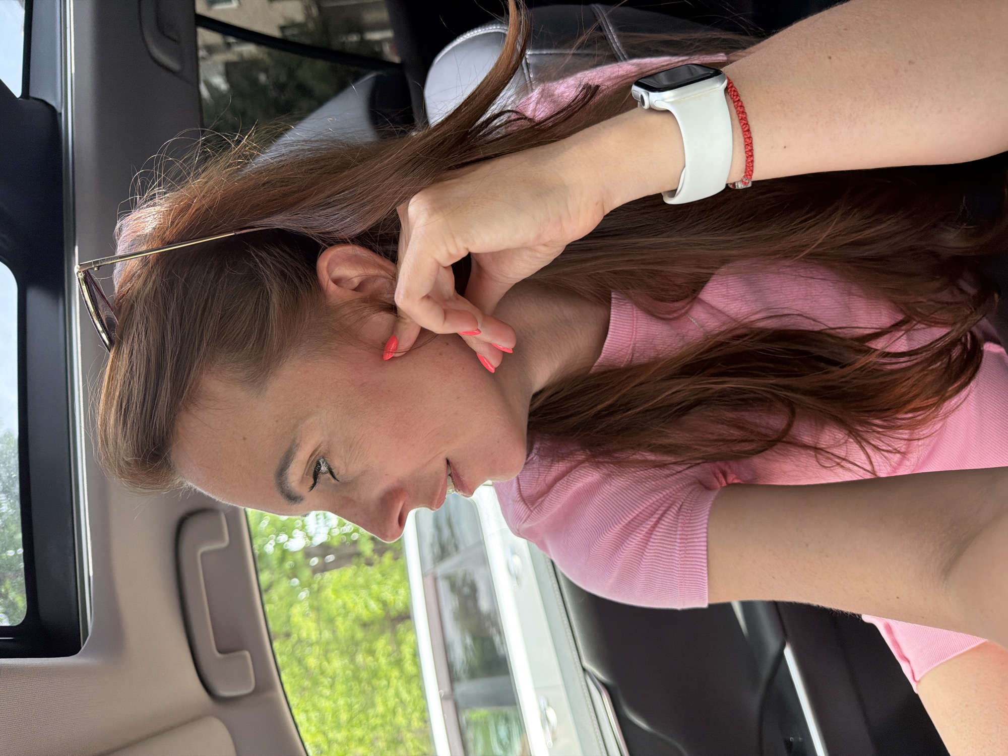 A woman with long, reddish-brown hair and bright pink fingernails is sitting in the passenger seat of a car, looking out the window. She is wearing a pink t-shirt and a white smartwatch with a red bracelet. Sunglasses are perched on top of her head. The car's sunroof is open, revealing a blurry green and white background suggesting trees and buildings. The scene appears to be in Birobidzhan, Russia.
