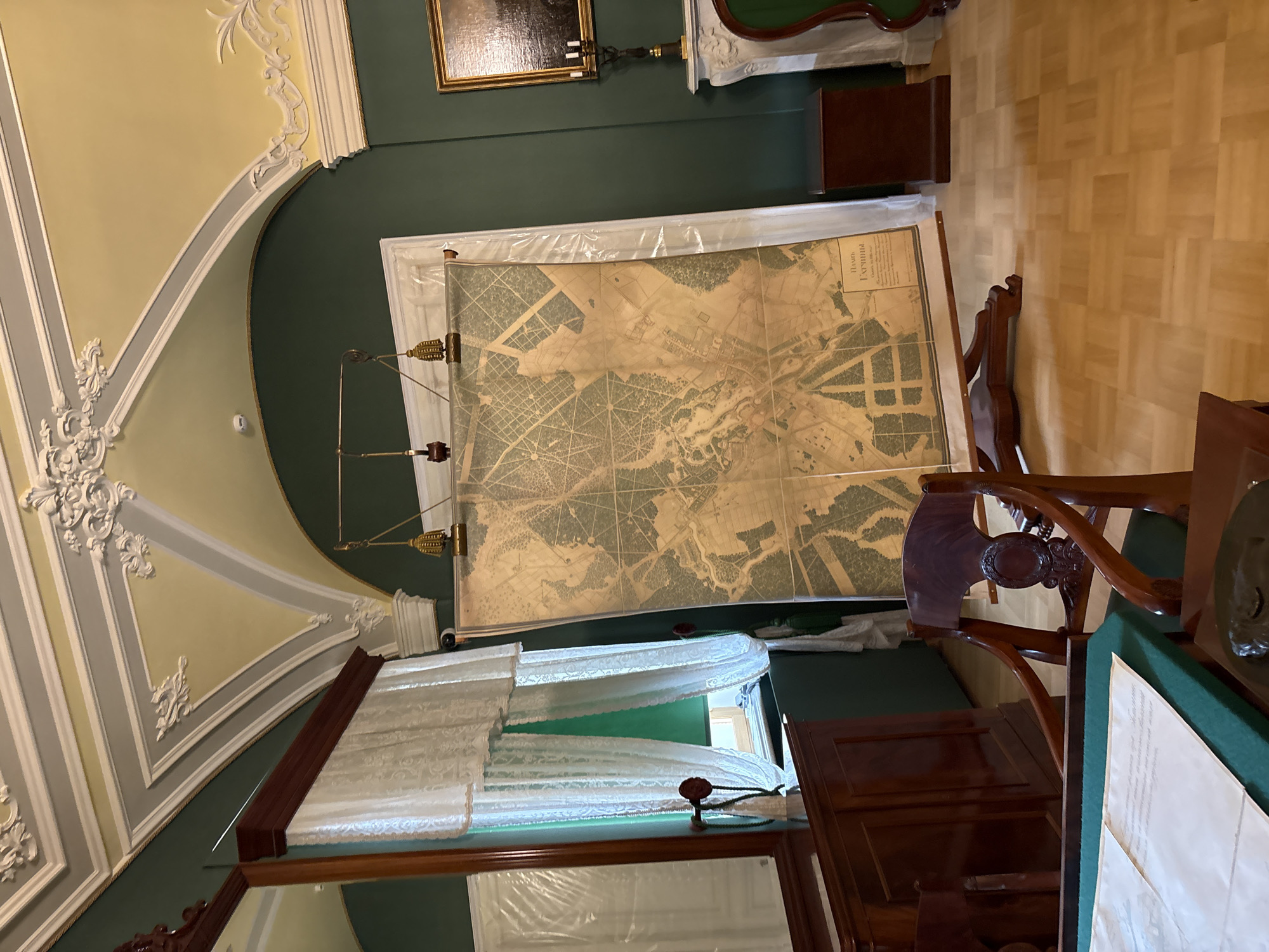 The image depicts an indoor scene, likely a museum or historical room, featuring a large antique map displayed on a wooden easel. The map is positioned in the center of the frame, leaning against a dark green wall. The map itself appears to be a detailed topographic or city plan, rendered in muted earth tones, with intricate lines and markings. The top of the map is secured by clips to a horizontal rod, which is suspended by wires and a decorative gilded finial.
To the left of the map, a tall, dark wooden cabinet with a mirror is visible. White lace curtains hang over a window behind this cabinet, and a green fabric is visible underneath. A dark wooden desk with papers on it is in the foreground, partially obscuring a dark wooden chair with an ornate backrest. To the right of the map, a white fireplace mantel is present, adorned with a framed painting above it and a candelabra. A small, dark wooden cabinet sits to the right of the fireplace. The room has a parquet wooden floor. The ceiling is arched and painted in shades of yellow and white, with ornate plasterwork.
The map has Russian text at the bottom, which reads "Планъ Гатчины" (Plan of Gatchina) and "съ обозначеніемъ улицъ и домовладеній" (with the designation of streets and properties). This indicates the location is Gatchina, Russia, and the map is a historical plan of the city. No people are visible in the image. The overall impression is one of a preserved historical interior, showcasing a significant cartographic artifact.