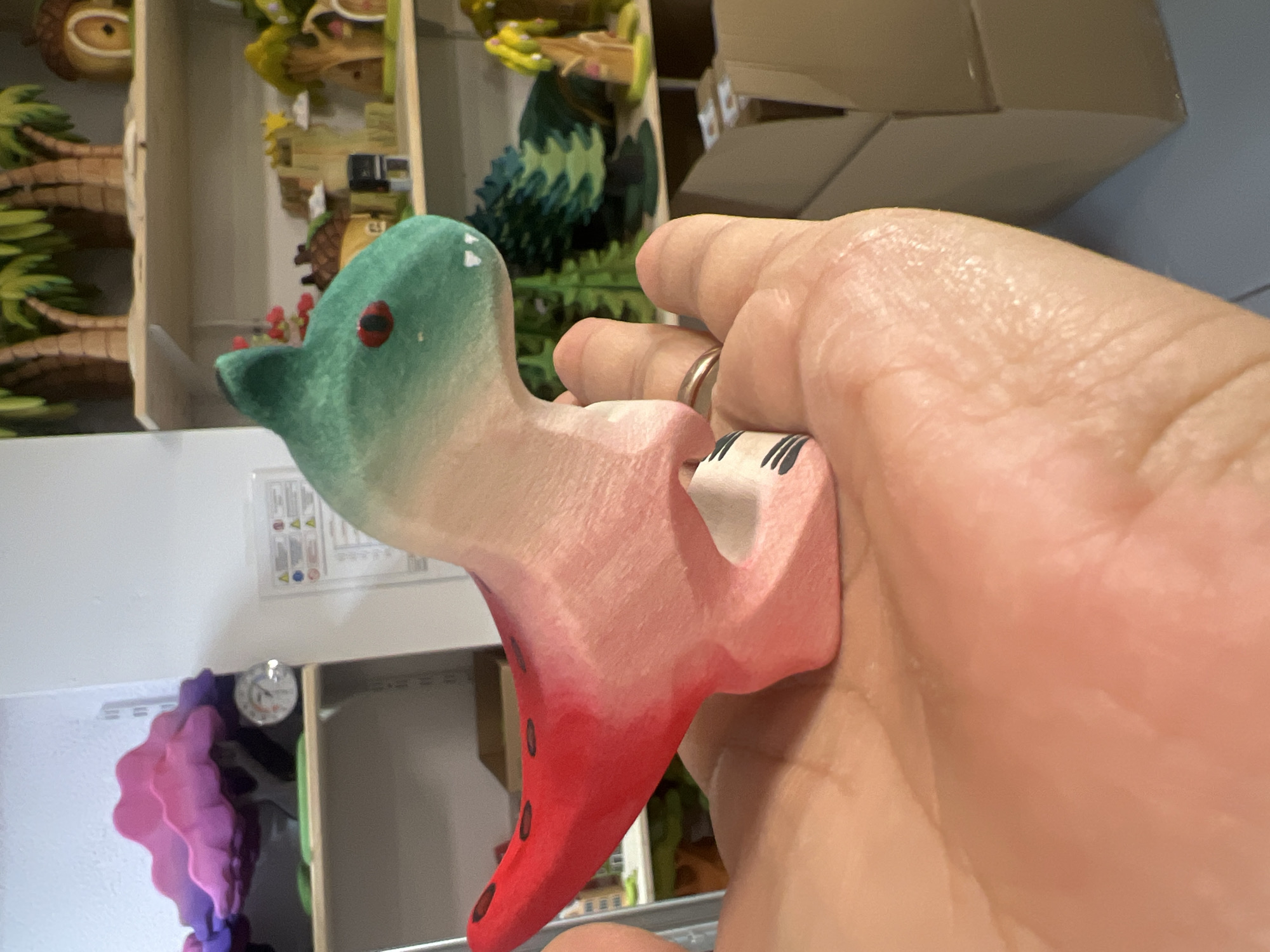 A person's hand is holding a wooden toy dinosaur that is predominantly red and green. The toy has black markings on its tail and a ring is visible on the person's finger. The background consists of shelves filled with various wooden toys, including trees and houses, suggesting a shop or display area. The lighting is bright, and the overall scene is well-lit.