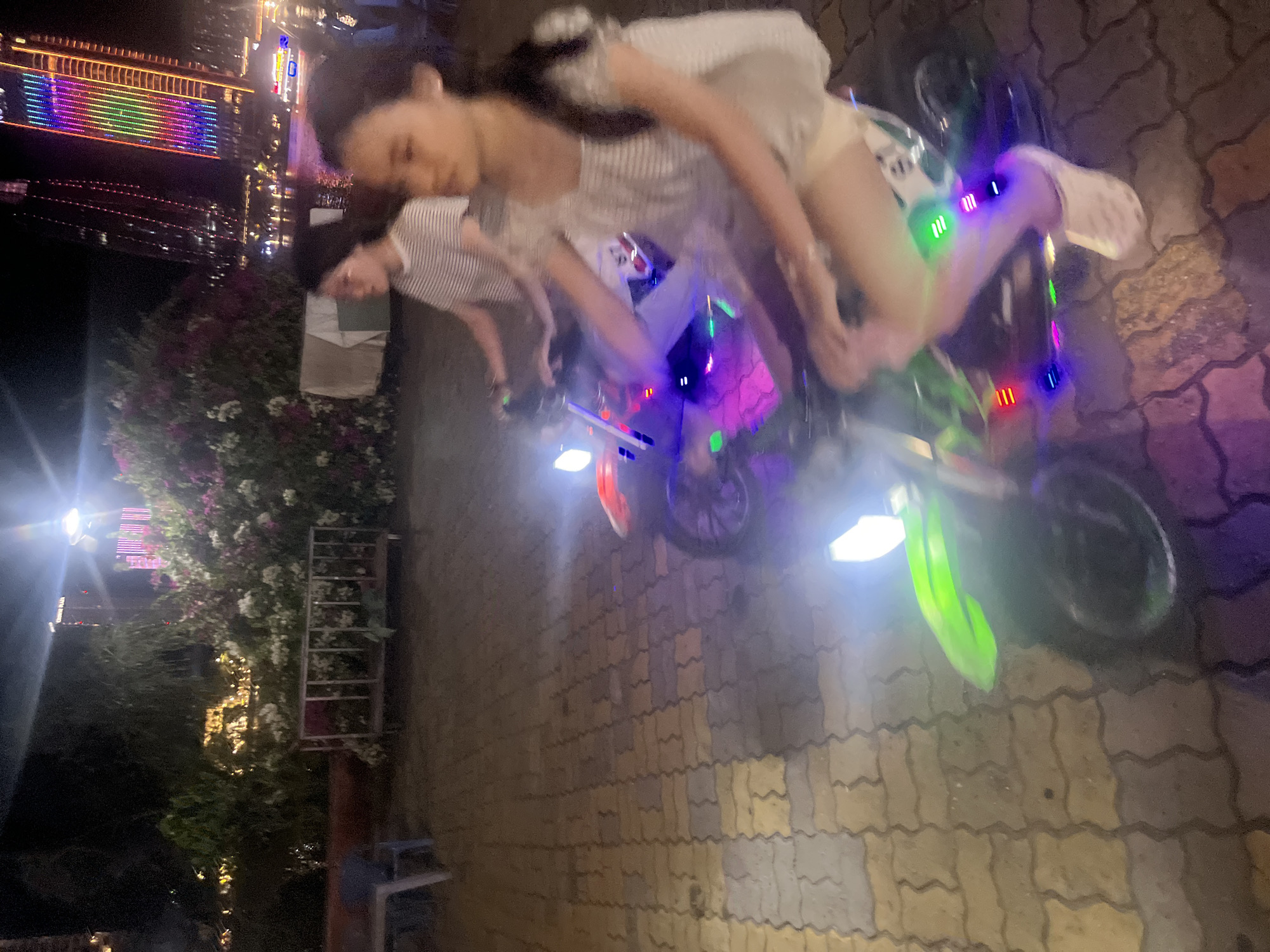 In Đà Nẵng, Vietnam, at night, two young females are riding miniature electric motorcycles along a paved walkway. The walkway is made of interlocking, multi-colored bricks. The female in the foreground, closer to the viewer, is wearing a white and striped top and beige shorts. She has dark hair tied back and is wearing white clogs. The motorcycle she is riding is decorated with numerous colorful LED lights. The second female, slightly further back and out of focus, is wearing a white and striped t-shirt and light-colored pants. The background features a lush display of flowering bushes with pink and white blooms, illuminated by artificial lights. Behind the foliage, a tall building is visible, adorned with a vibrant, multi-colored LED light display. Other structures and lights can be seen further in the distance, suggesting an urban waterfront setting. The scene is captured in low light conditions, with the motorcycle lights and distant building lights being the primary light sources.