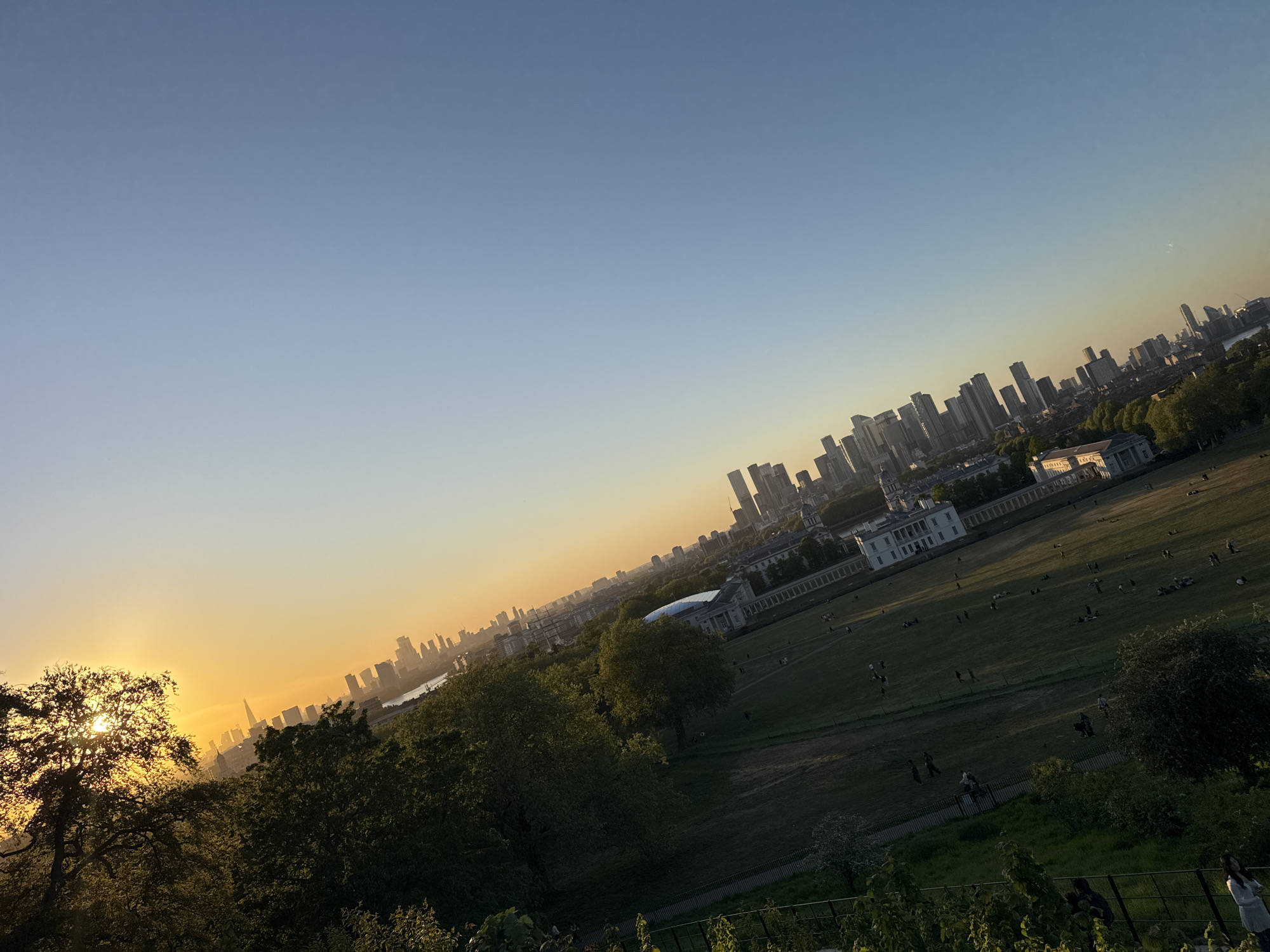 This media file presents a panoramic landscape view, likely captured from an elevated position such as a hill, looking out over an urban area during sunset. The golden sun is partially visible on the far left, creating a warm, intense glow that transitions across the sky from bright orange and yellow near the horizon to a clear, soft blue overhead. This indicates either sunrise or sunset, with the long shadows suggesting the latter.
In the foreground and midground, a vast expanse of green parkland dominates, with numerous small figures of people scattered across the grass and walking along pathways. They appear to be enjoying leisure activities, relaxing or strolling. Several mature trees are visible, some silhouetted against the bright sky. A prominent white, classical-style building with columns is situated in the mid-distance, surrounded by more greenery, and beyond it, a cluster of modern city buildings rise.
The background features a dense urban skyline, characteristic of a major city. Many tall buildings and skyscrapers are silhouetted against the fading light, with what appears to be a river winding through the city. The architecture strongly suggests the London skyline, with the cluster of buildings resembling Canary Wharf to the right, and other central London landmarks further left. Given the location context, this view is consistent with Greenwich Park looking towards the Thames and the city beyond.
The overall scene conveys a sense of tranquility and an appreciation for the urban landscape bathed in the warm light of the setting sun. There is no visible text in the image.