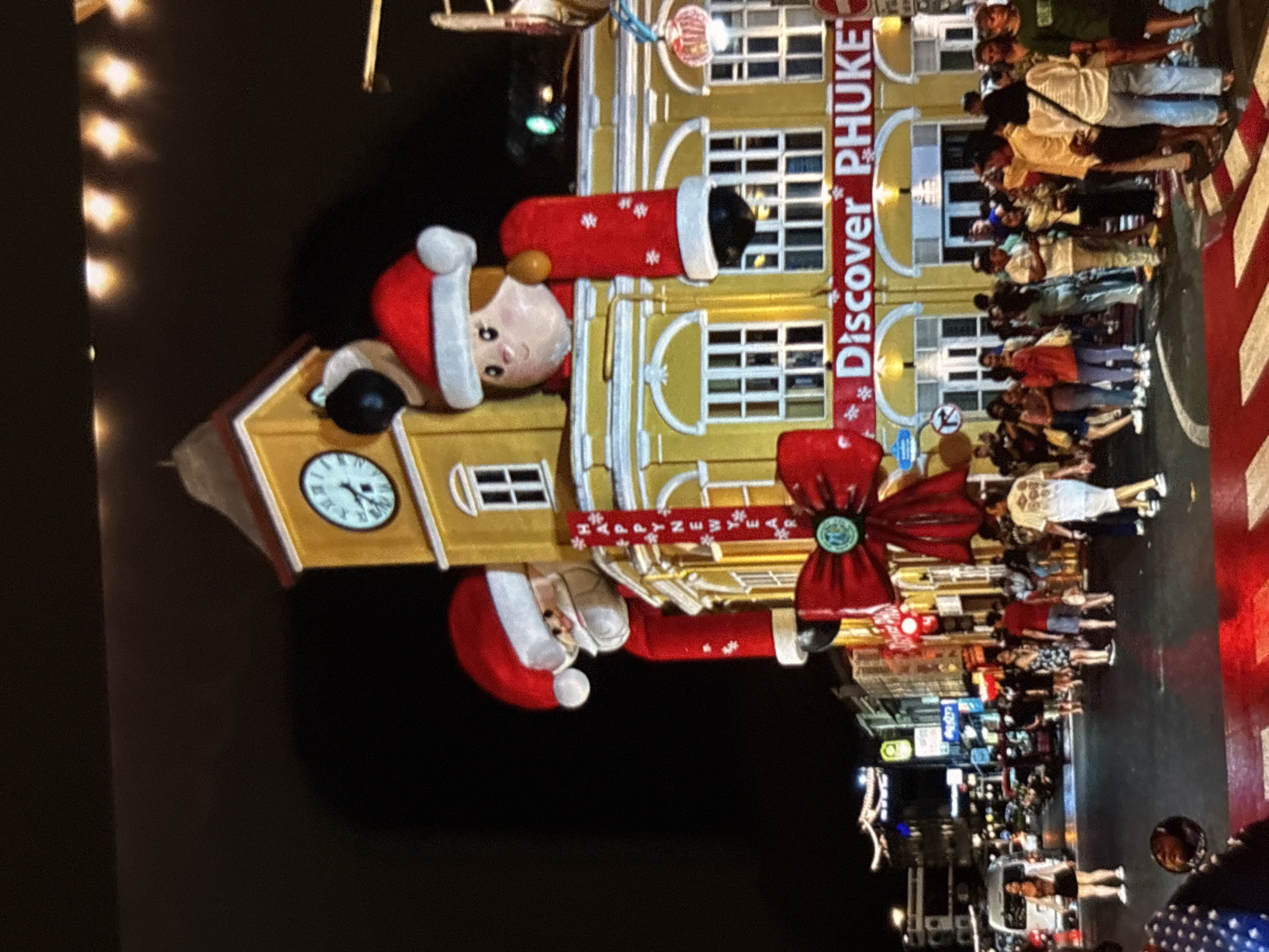 At night in Chalong, Thailand, a yellow colonial-style building is adorned with festive Christmas decorations. A large inflatable Santa Claus and a festive female figure, both wearing red Santa hats, are perched on top of the building, appearing to peek over the edge. The building itself features arched windows and a prominent clock tower. A red banner proclaiming "Discover PHUKET" is stretched across the facade. Below, a bustling street scene unfolds with a crowd of people walking and standing on the sidewalk and crossing the street. Vehicles, including a white van and motorbikes, are visible in the background. The street is lit by overhead lights and illuminated storefronts, creating a vibrant and lively atmosphere.