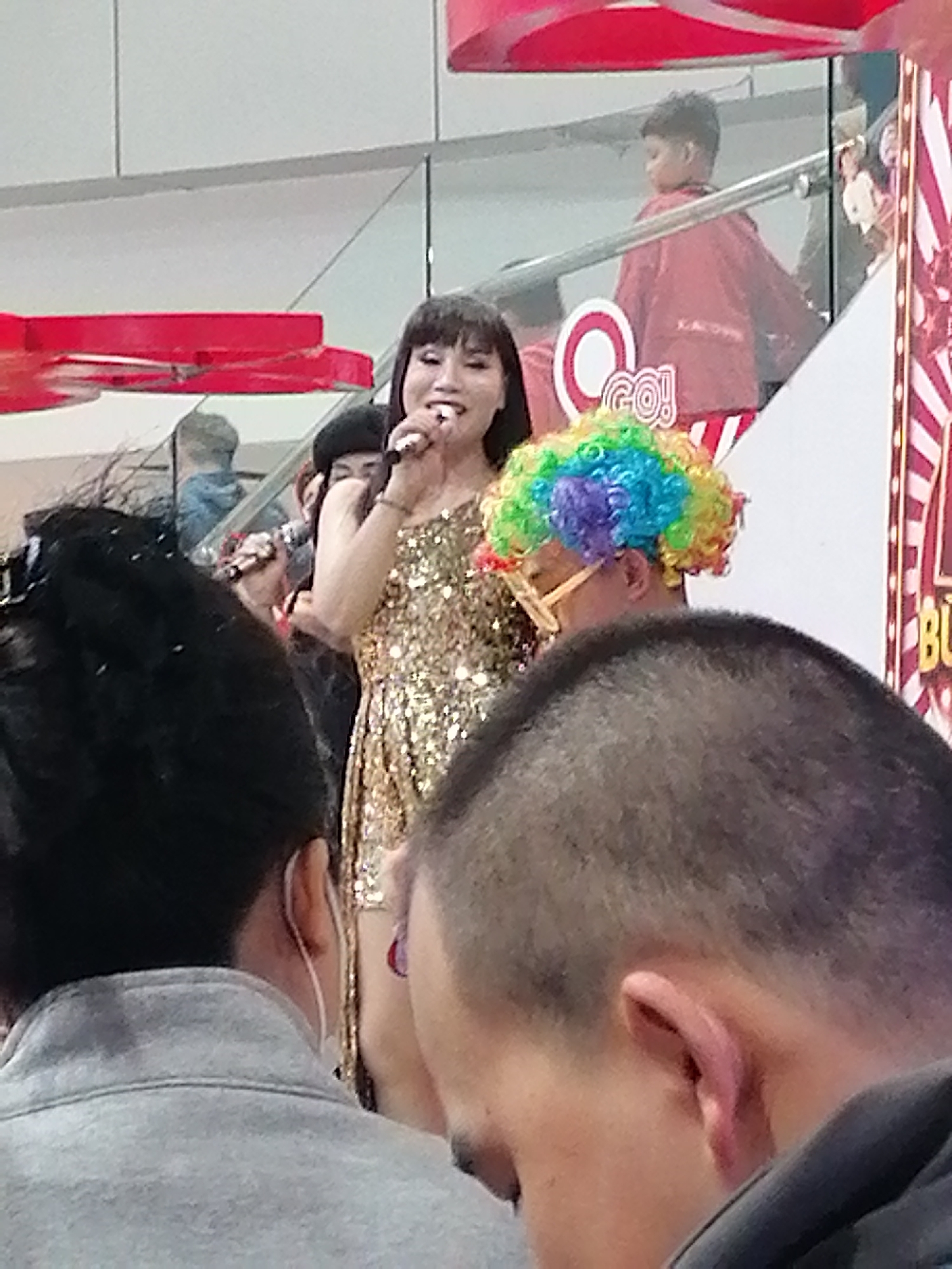 The image captures a lively scene at an indoor event, possibly a mall or entertainment venue, in Da Lat, Vietnam. A woman in a sparkling gold sequined dress is singing into a microphone, her expression one of joy. She has dark hair and is wearing elaborate eye makeup. To her right, a person sporting a vibrant rainbow-colored wig and oversized glasses is partially visible, suggesting a festive or performative atmosphere. In the background, other individuals can be seen, including a young boy in a red jacket and people standing on an elevated walkway with a railing. The environment appears to be a multi-level structure with a modern architectural design, indicated by the white walls and railings. Red decorative elements, possibly part of a stage setup or signage, are also present. The focus is on the performers and the engaged audience, creating a sense of public celebration.